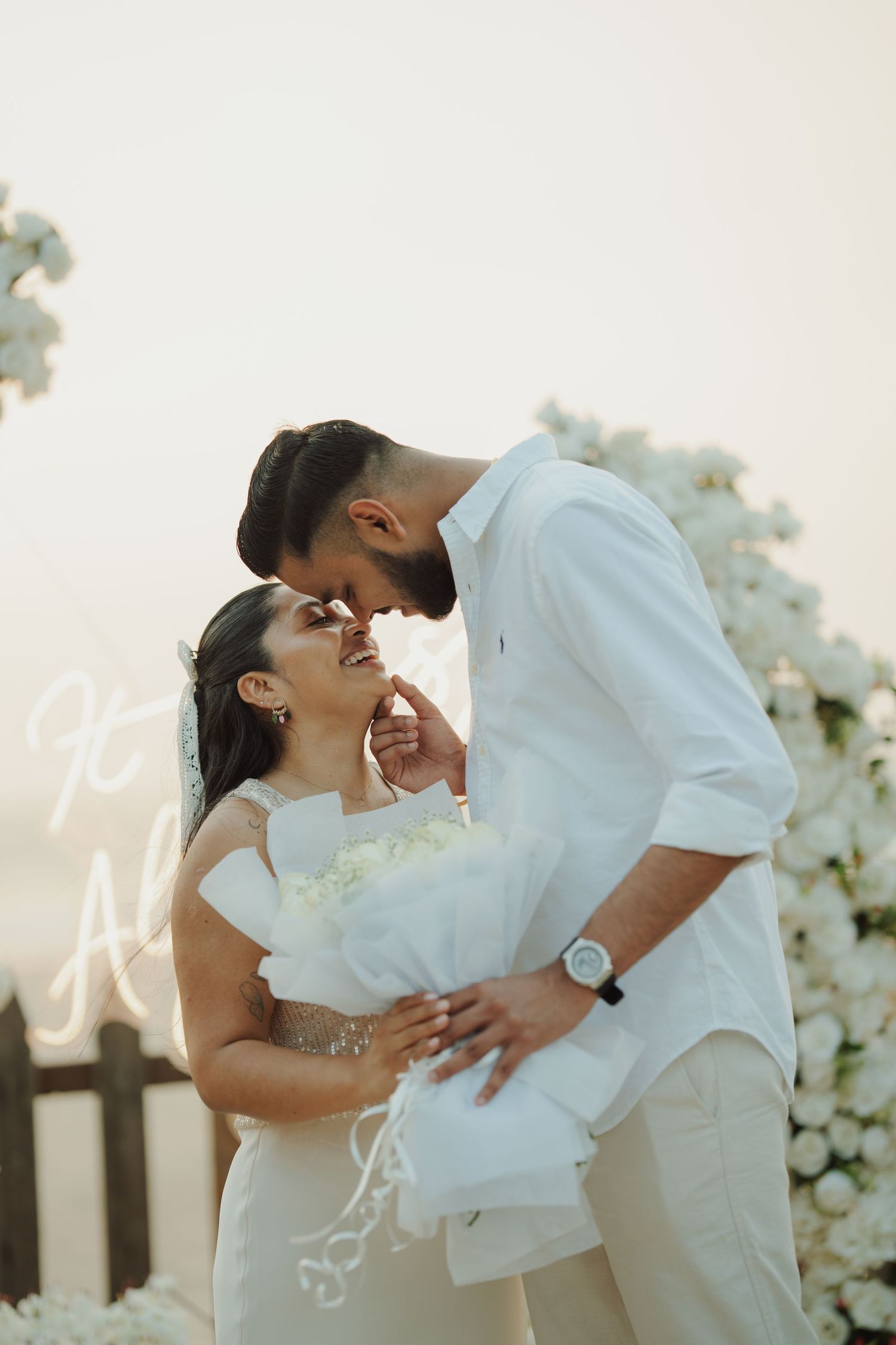 Couple in white attire sharing a romantic moment at an outdoor wedding ceremony with white floral decorations.