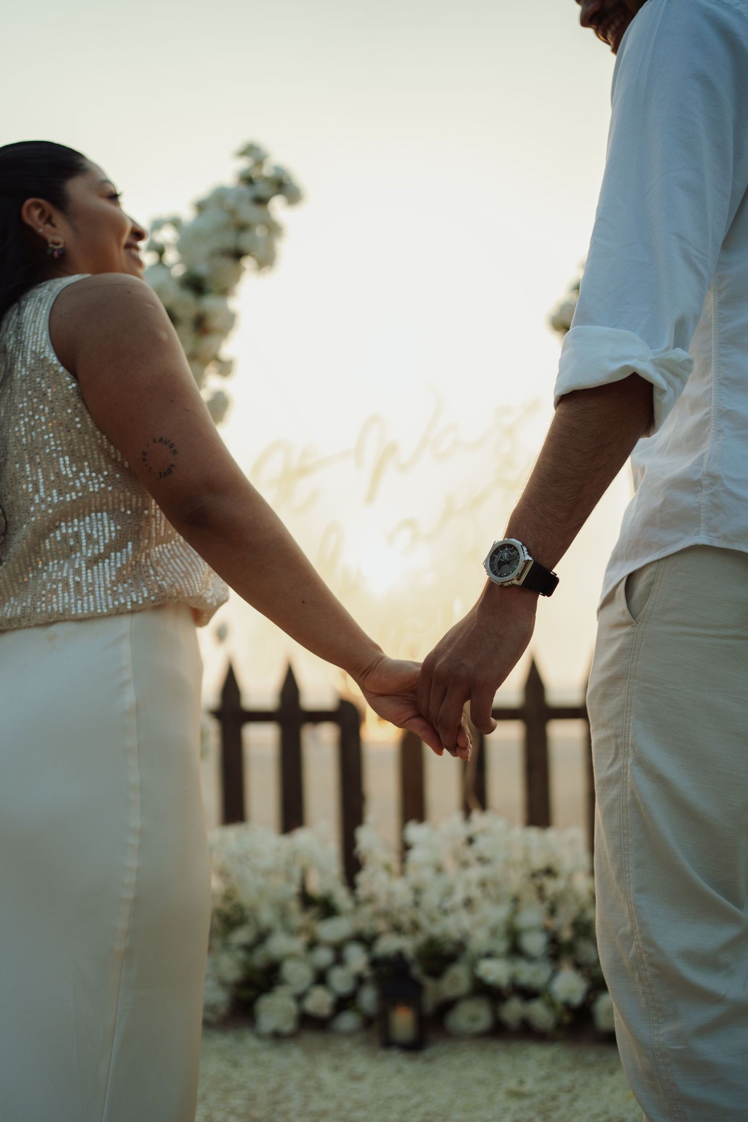 Couple holding hands at sunset wedding ceremony with white floral arrangements and wooden fence backdrop.
