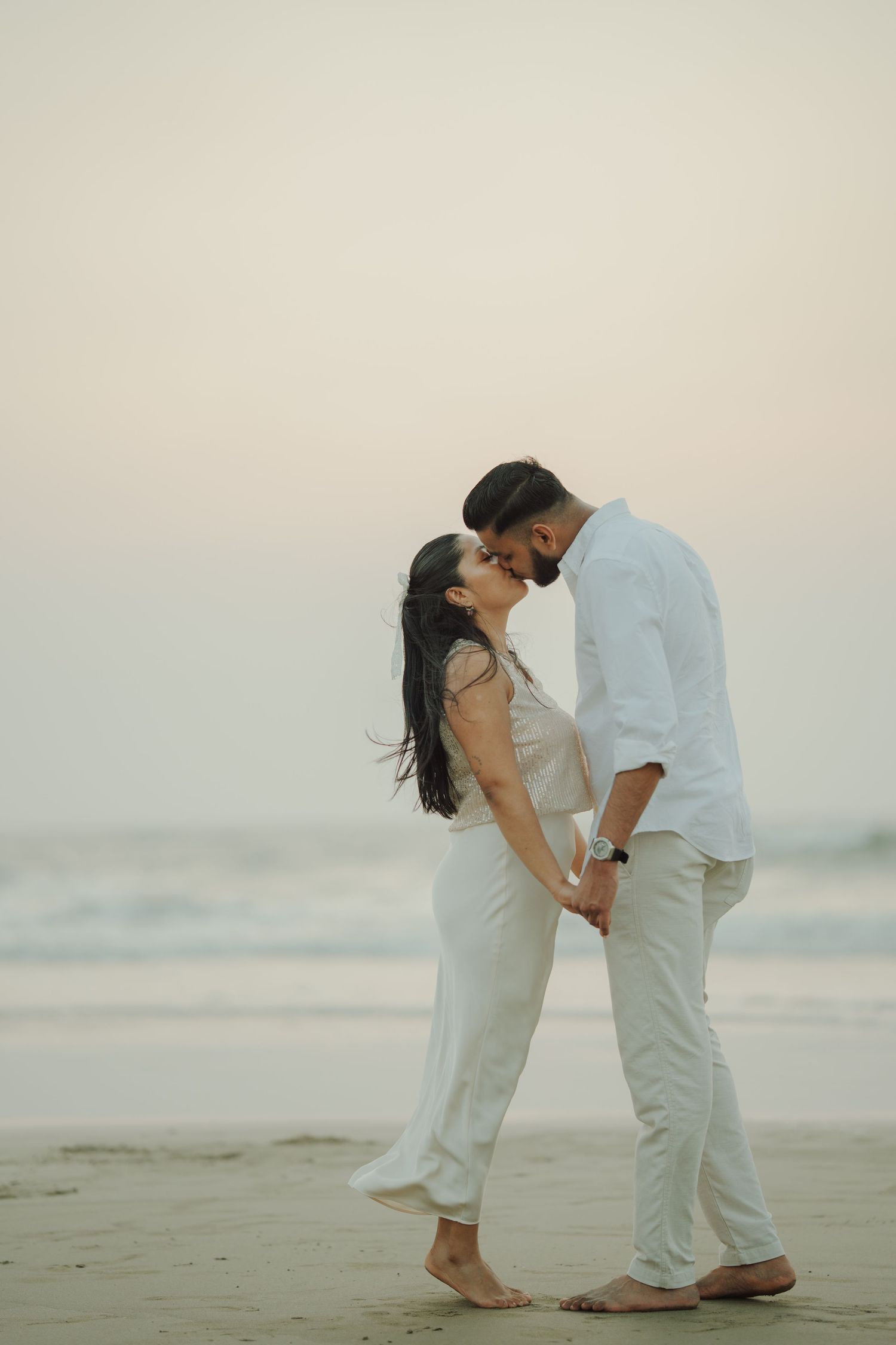 Couple in white outfits sharing a romantic moment on a beach at sunset, holding hands barefoot on the sand.