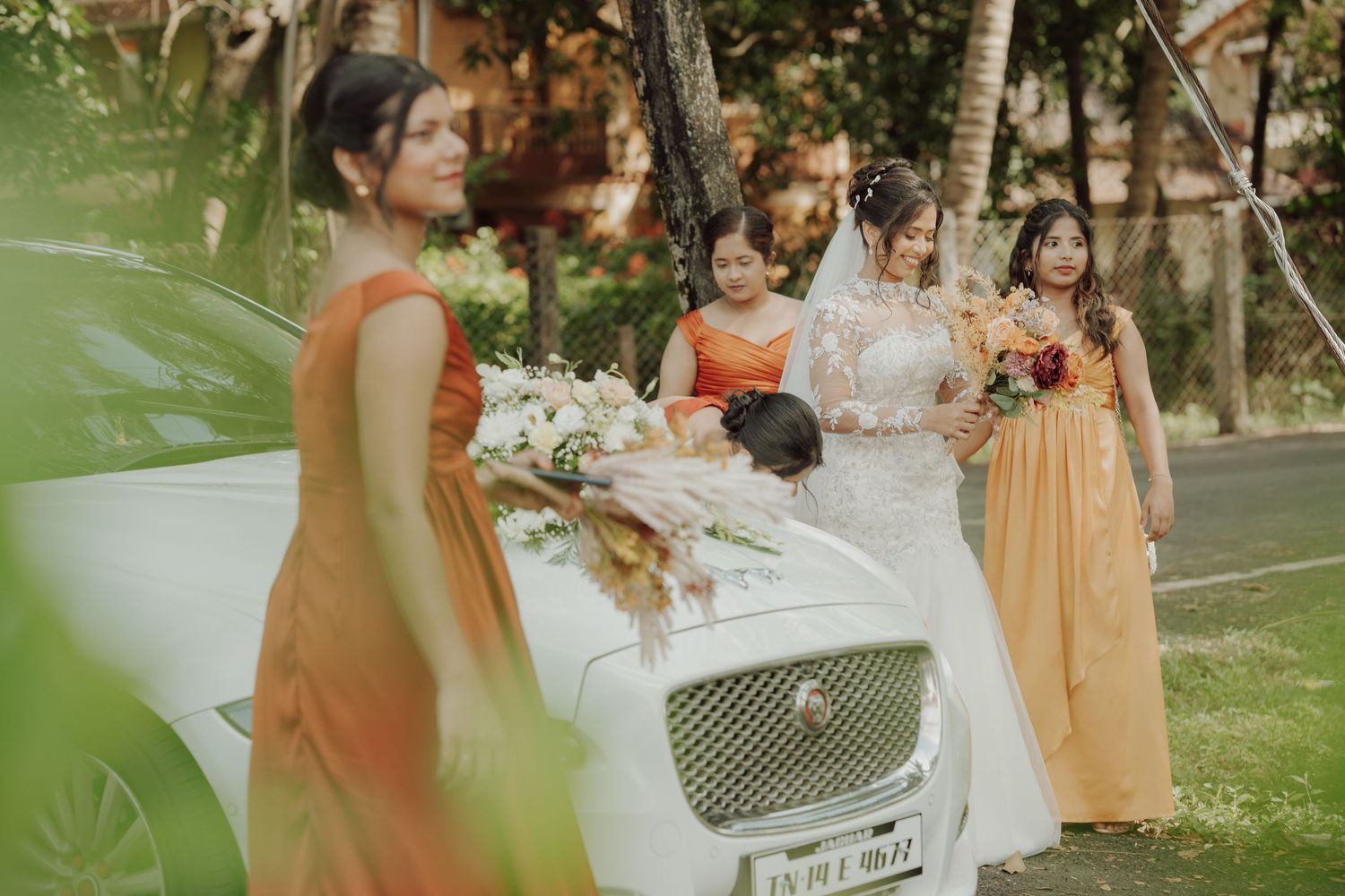 Bride in a white veil beside a white luxury car decorated with flowers, with bridesmaids in orange dresses.