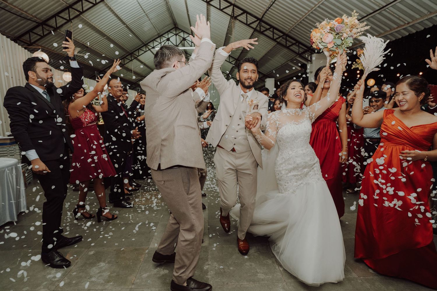 Bride and groom enter their wedding reception as guests cheer and throw confetti petals in a festive covered venue.