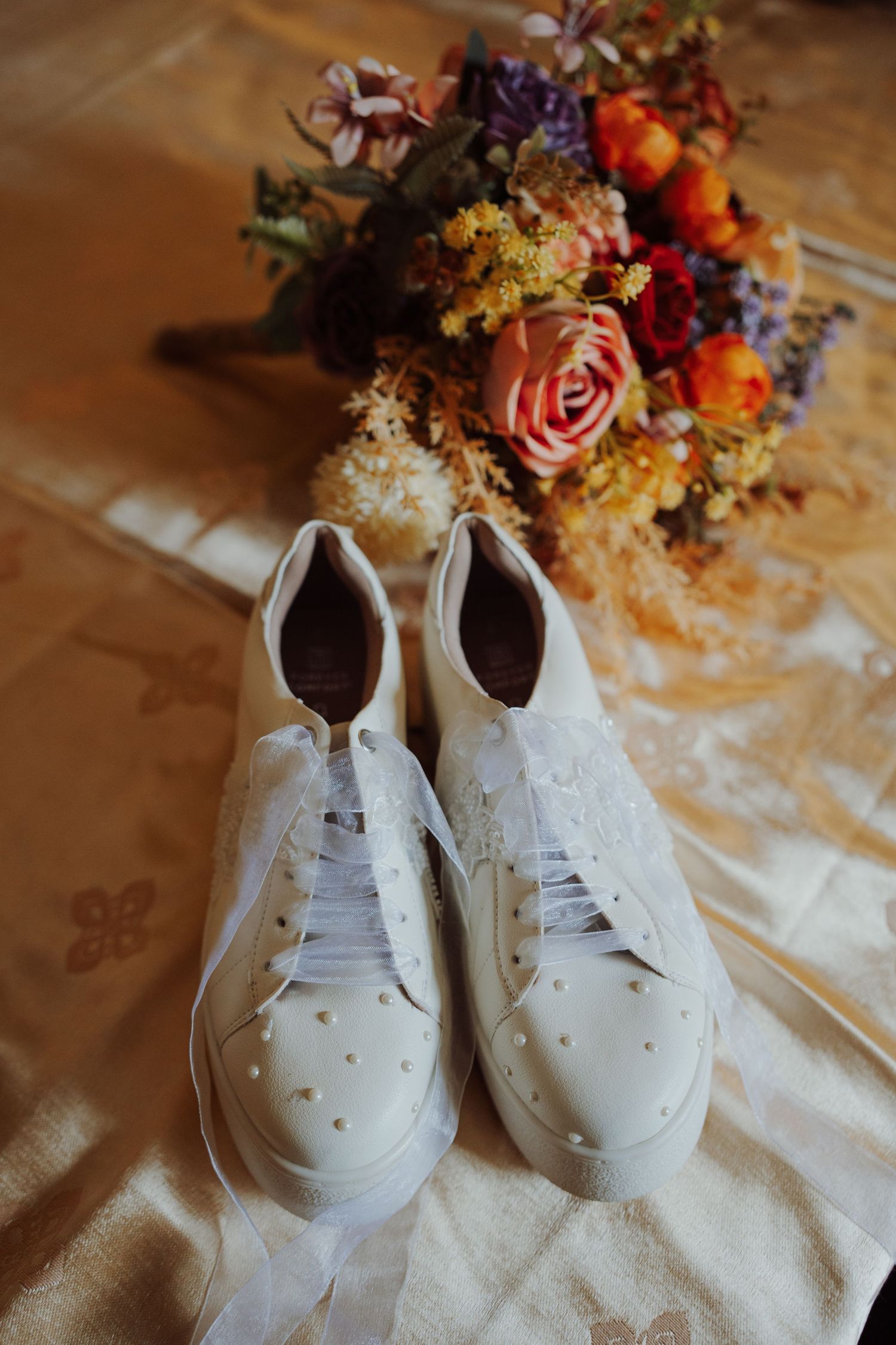 White beaded sneakers with sheer ribbon laces beside a colorful bridal bouquet of roses and wildflowers on gold fabric.