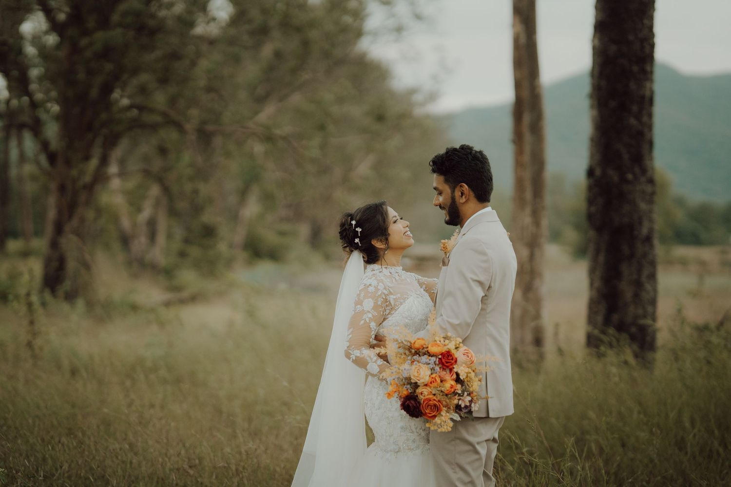 Bride in a white lace gown and veil holding an orange bouquet embraces her groom in a beige suit in a grassy forest clearing.
