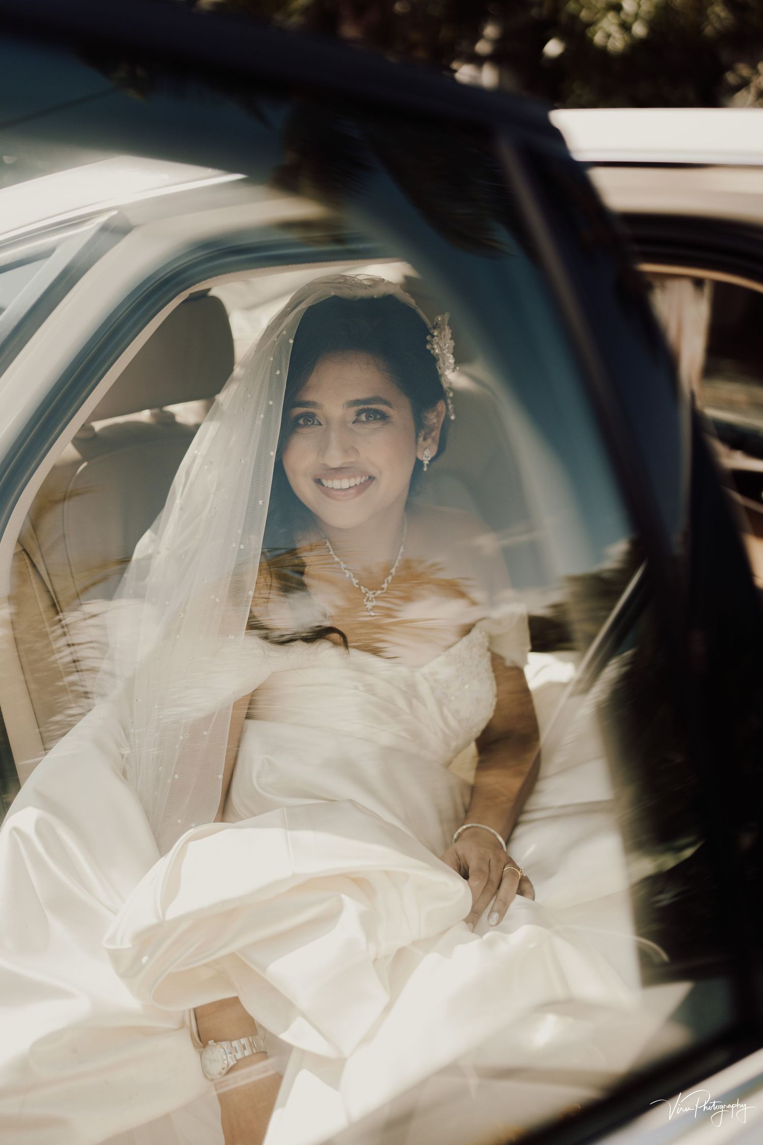 Through a car window, a bride wearing a white wedding dress and veil sits elegantly in the vehicle's back seat.