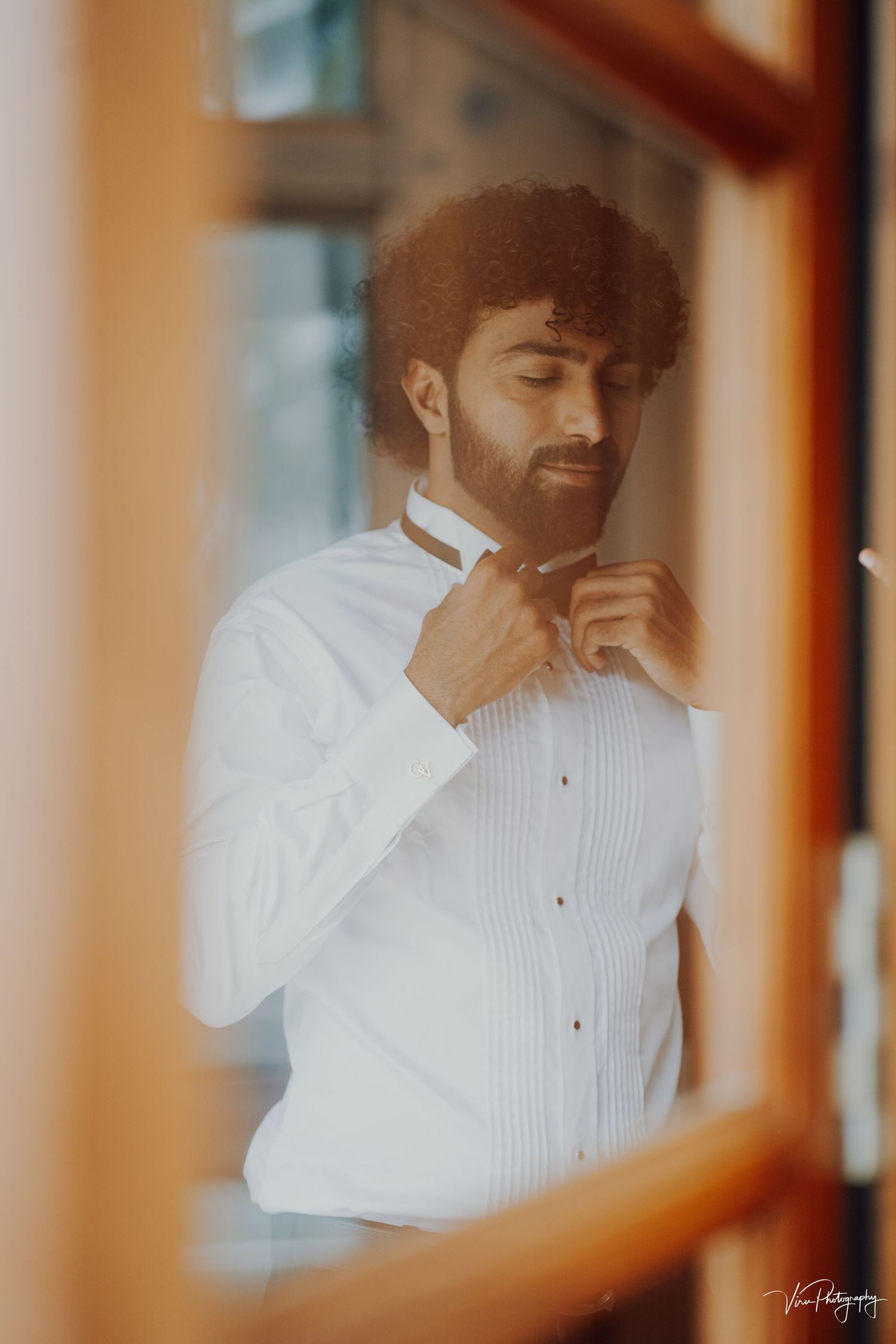 A person in a white formal dress shirt adjusts their bowtie while looking down with a gentle smile in warm lighting.