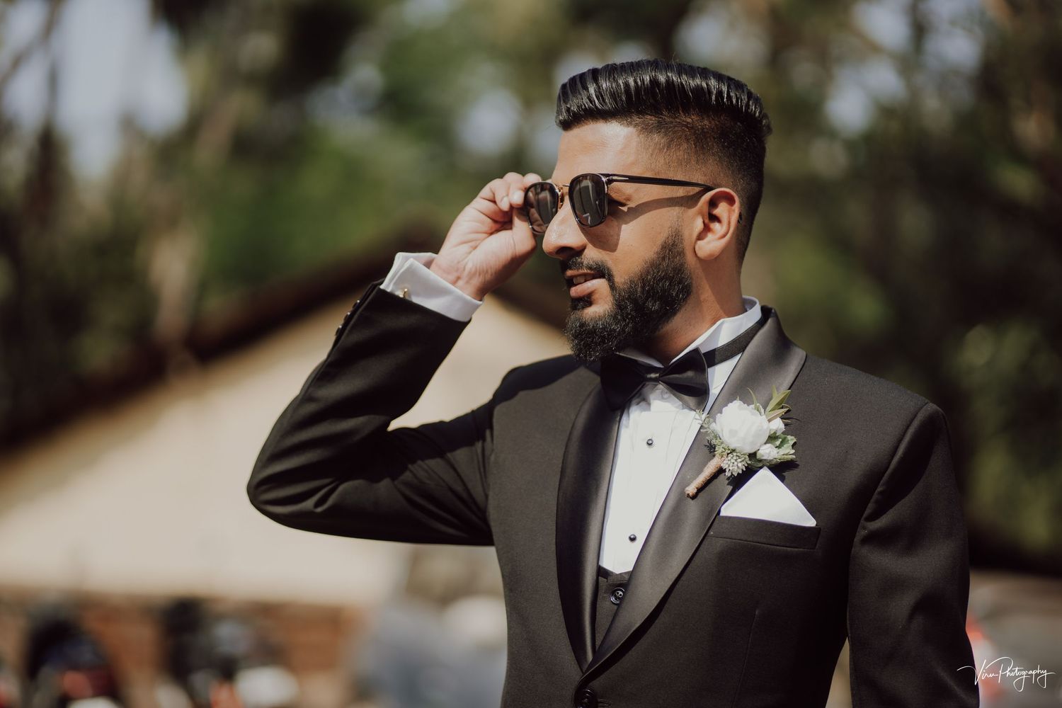 Groom in black tuxedo and bowtie adjusts sunglasses while posing outdoors for wedding photography.