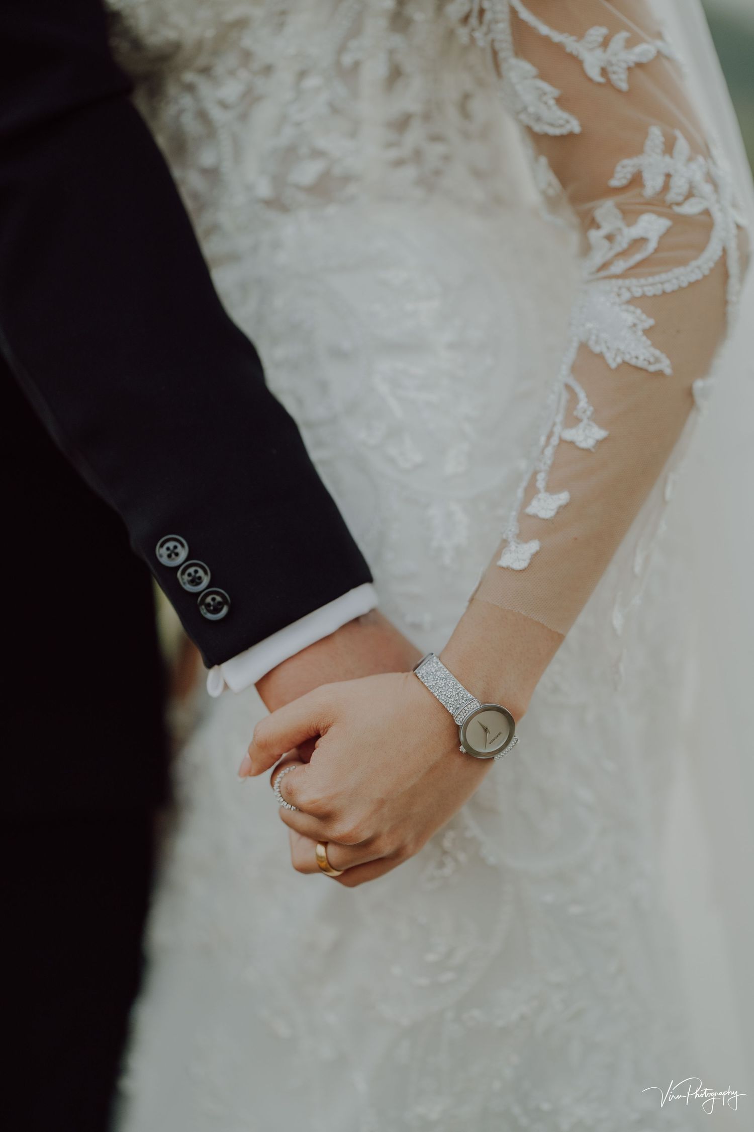 Close-up of intertwined hands showing delicate lace sleeve detail and wristwatch at a wedding ceremony.