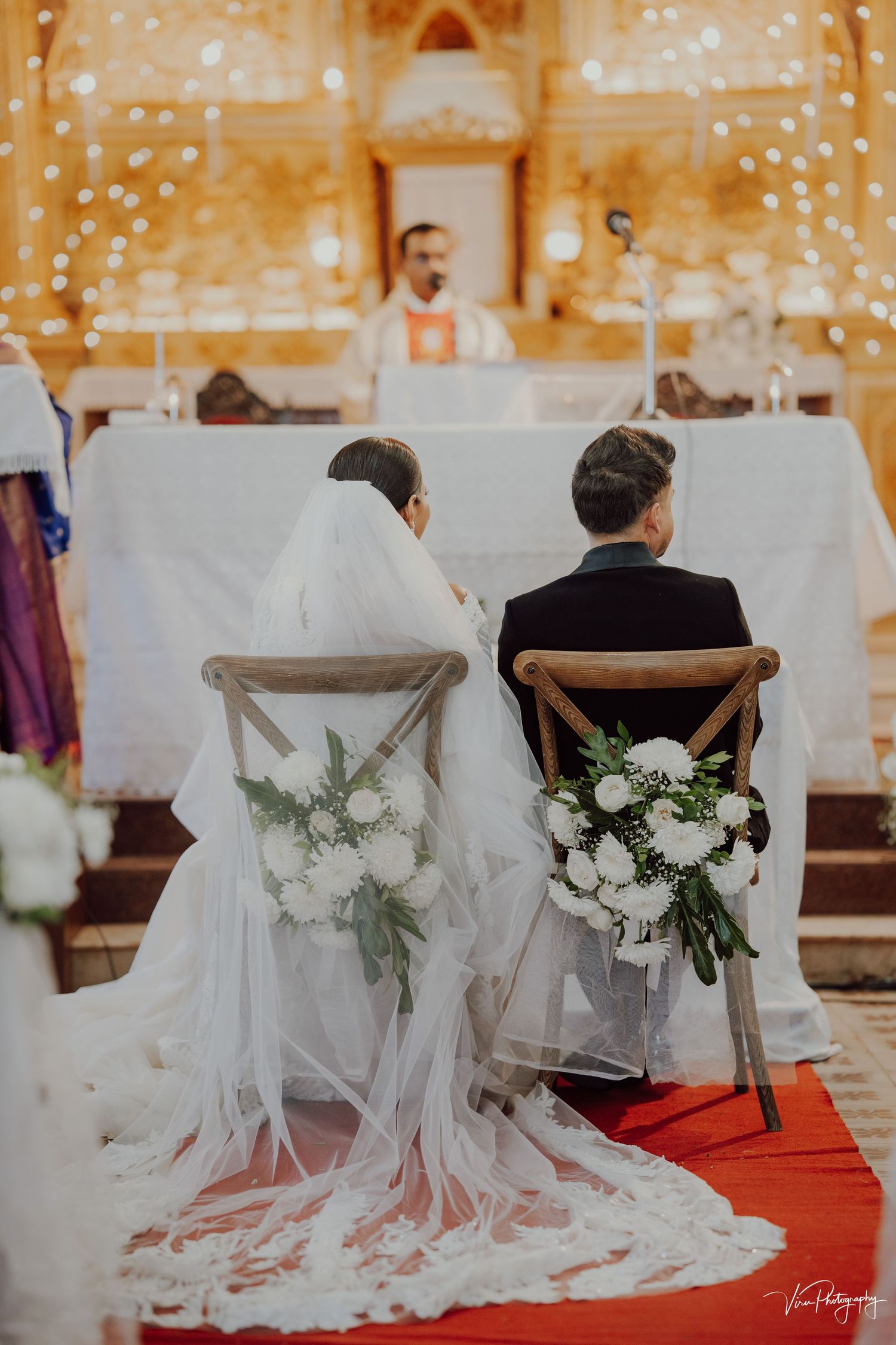 A couple sits during their wedding ceremony with white floral chair decorations and a flowing veil in a church setting.