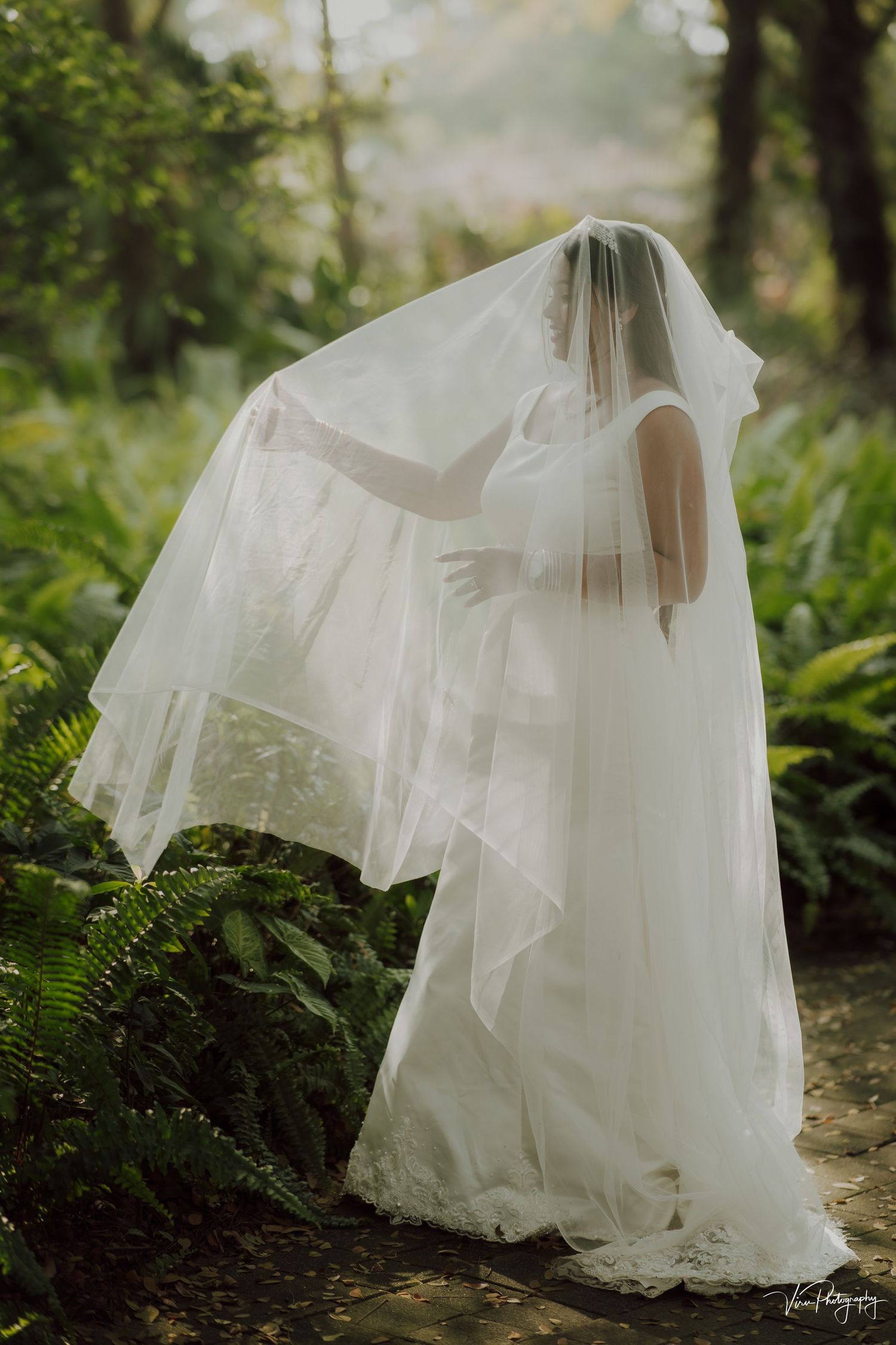A bride in a flowing white wedding dress and veil poses gracefully among lush ferns in a misty forest setting.