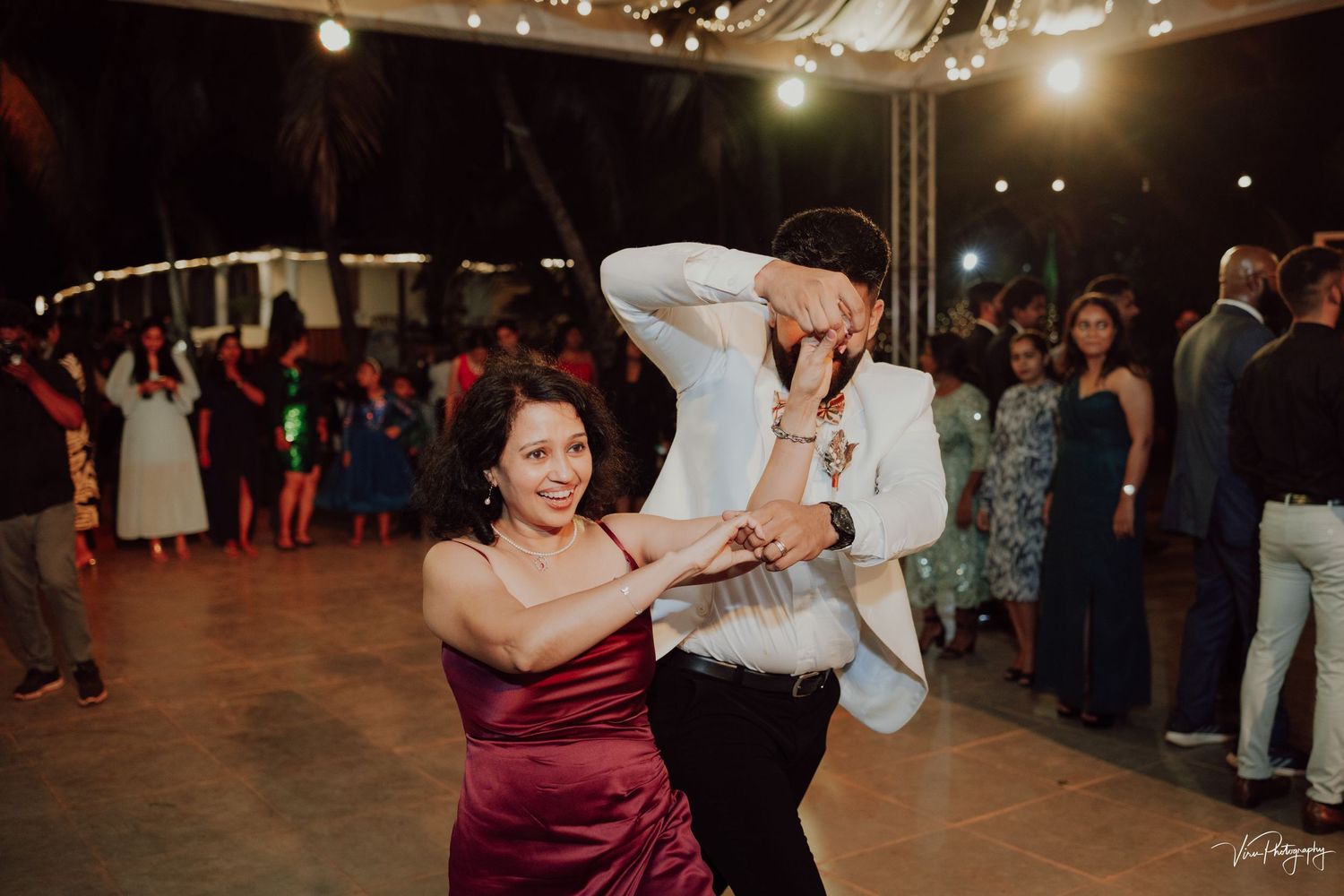 Two wedding guests dance energetically together on a reception dance floor with a crowd in the background.