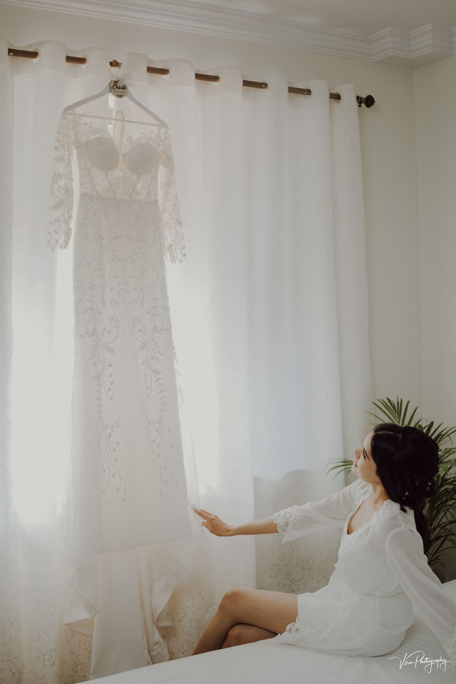 A delicate white lace wedding dress hangs from a curtain rod as someone in a white robe reaches toward it from a bed.