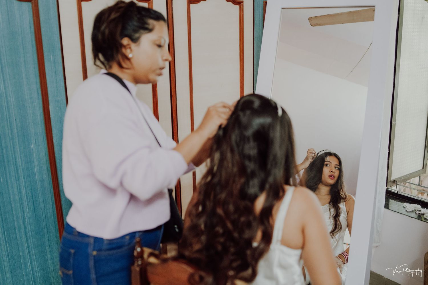 Stylist works on a client's long dark hair in a salon setting with mirrors and curtains visible.