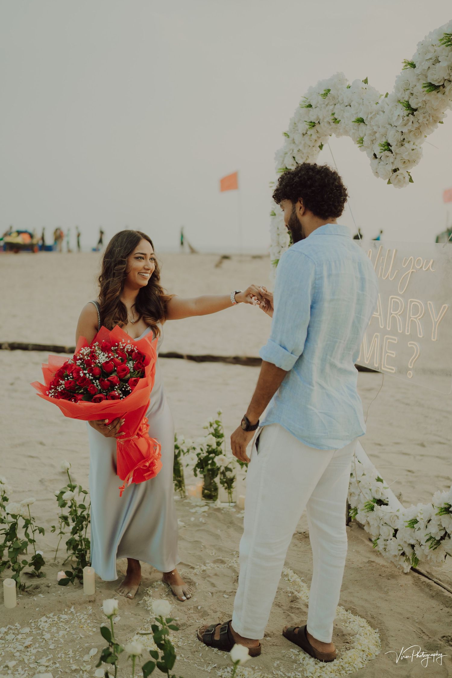 A romantic seaside proposal scene with a red bouquet of flowers and white floral decorations on the beach at sunset.