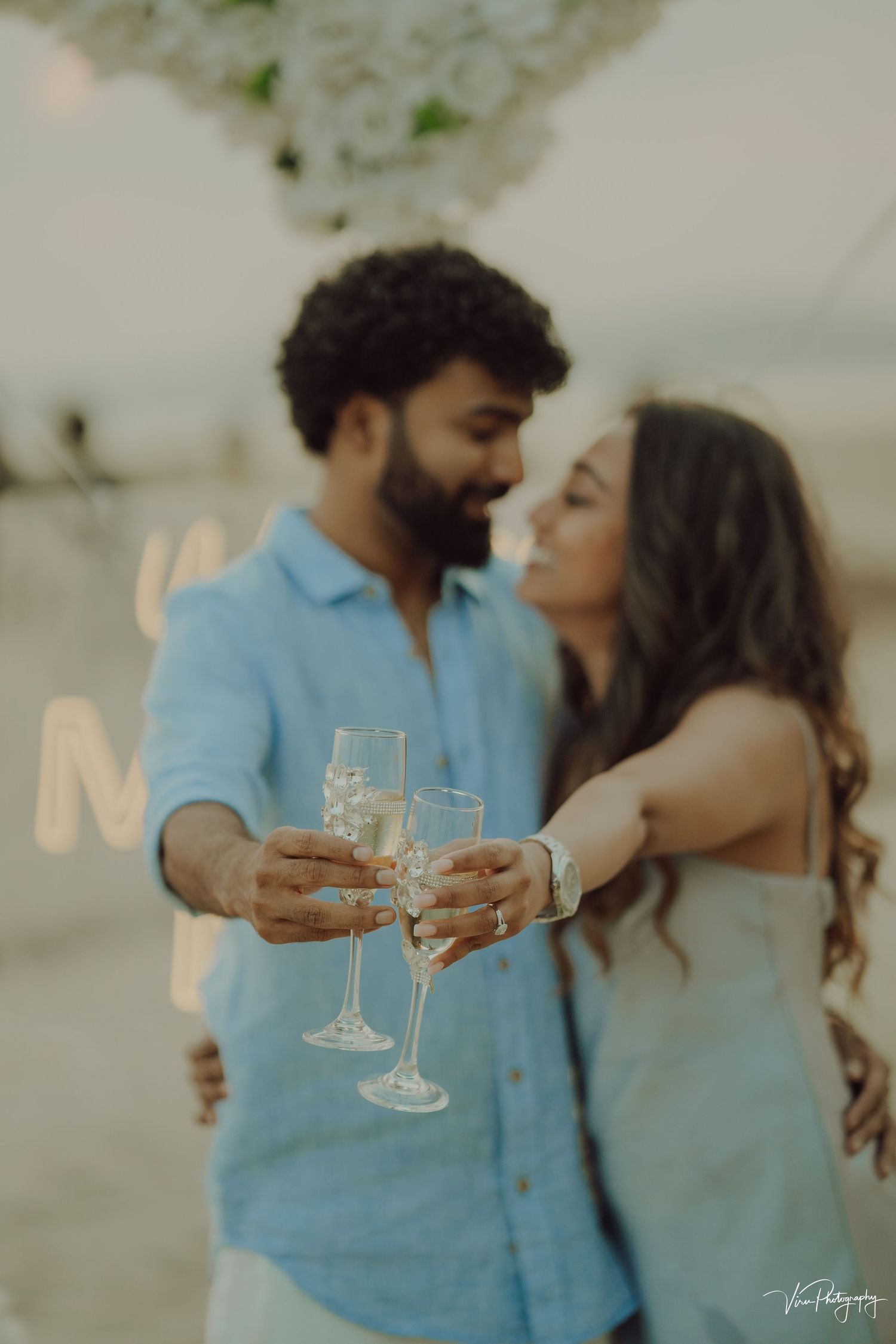 A couple in blue outfits share an intimate moment while toasting with champagne glasses at sunset.