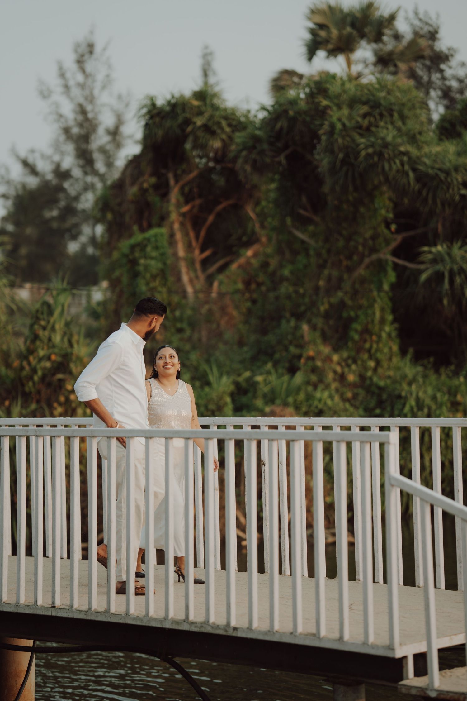 Couple in white outfits sharing a romantic moment on a white bridge surrounded by lush tropical greenery.