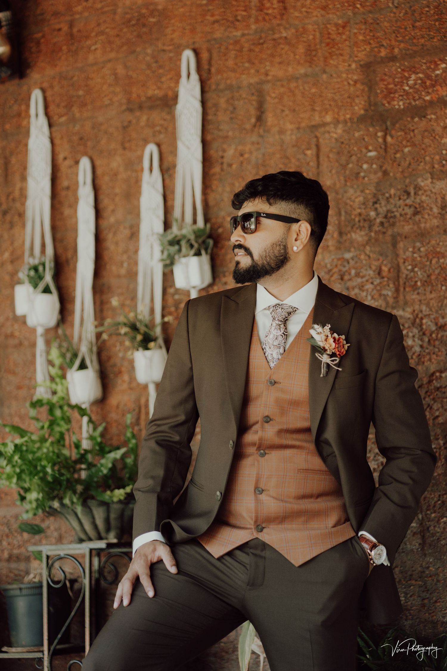Stylishly dressed groom in dark suit with tan vest poses against rustic brick wall with hanging potted plants.