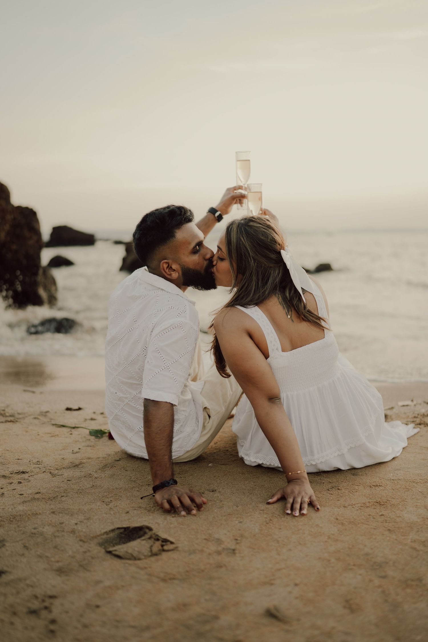 Romantic couple in white outfits lying together on a misty beach during sunset.