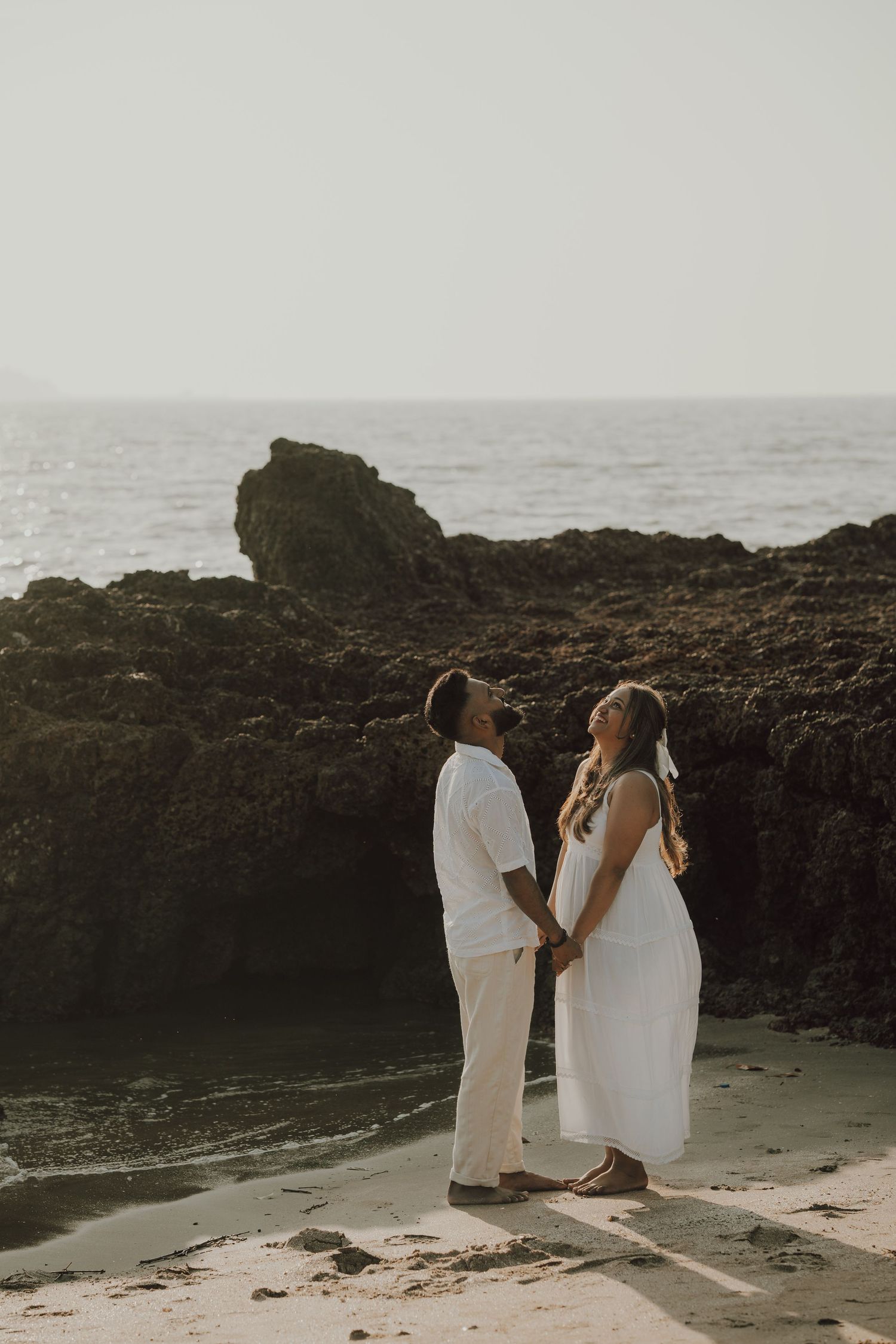 Two people in white outfits stand together on rocky coastal cliffs at sunset overlooking the ocean.