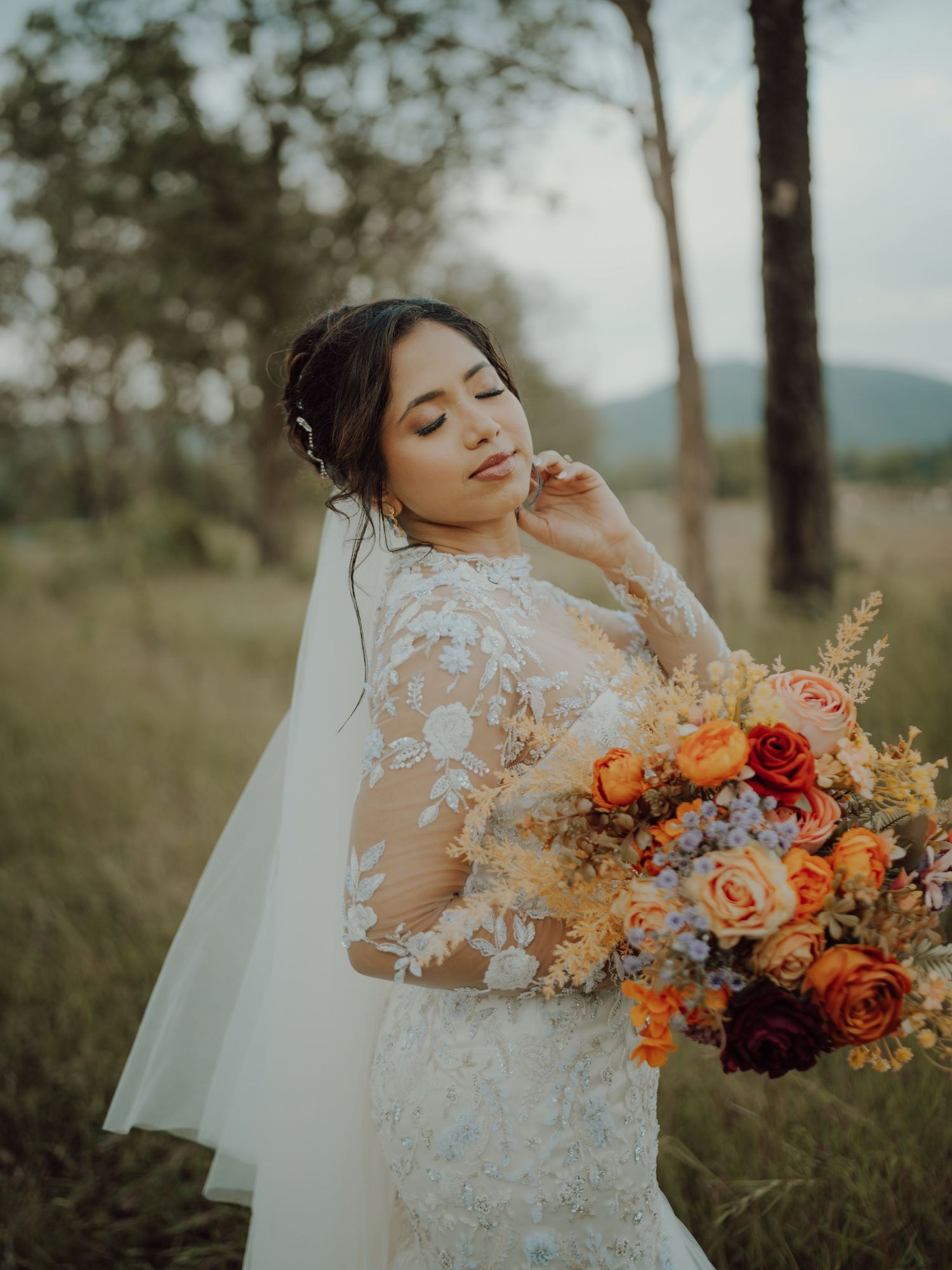 Bride in a lace wedding gown and veil poses in a grassy field holding a vibrant bouquet of orange and burgundy flowers.