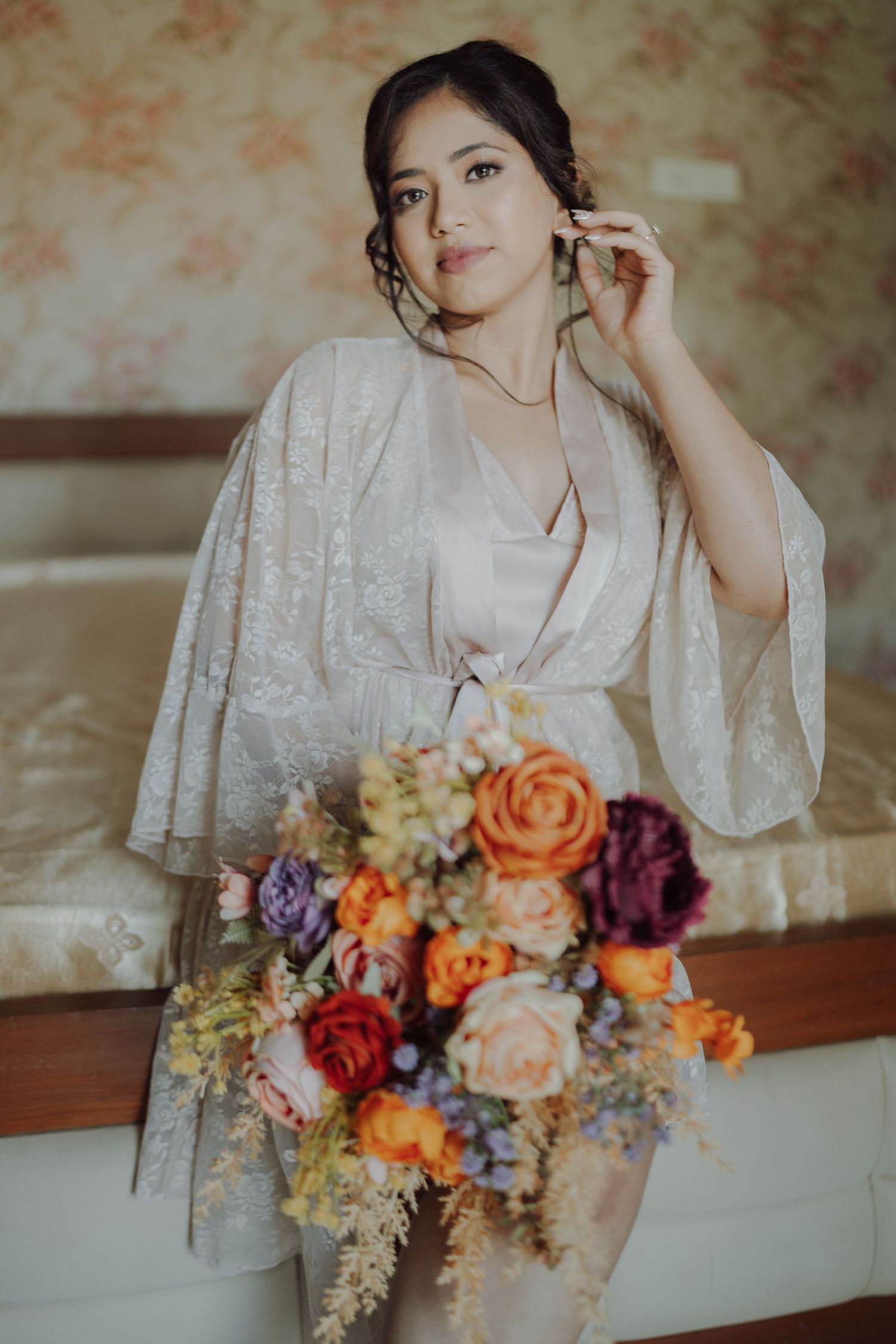 Bride in a satin and lace robe touches her earring while holding an orange and burgundy floral bouquet.