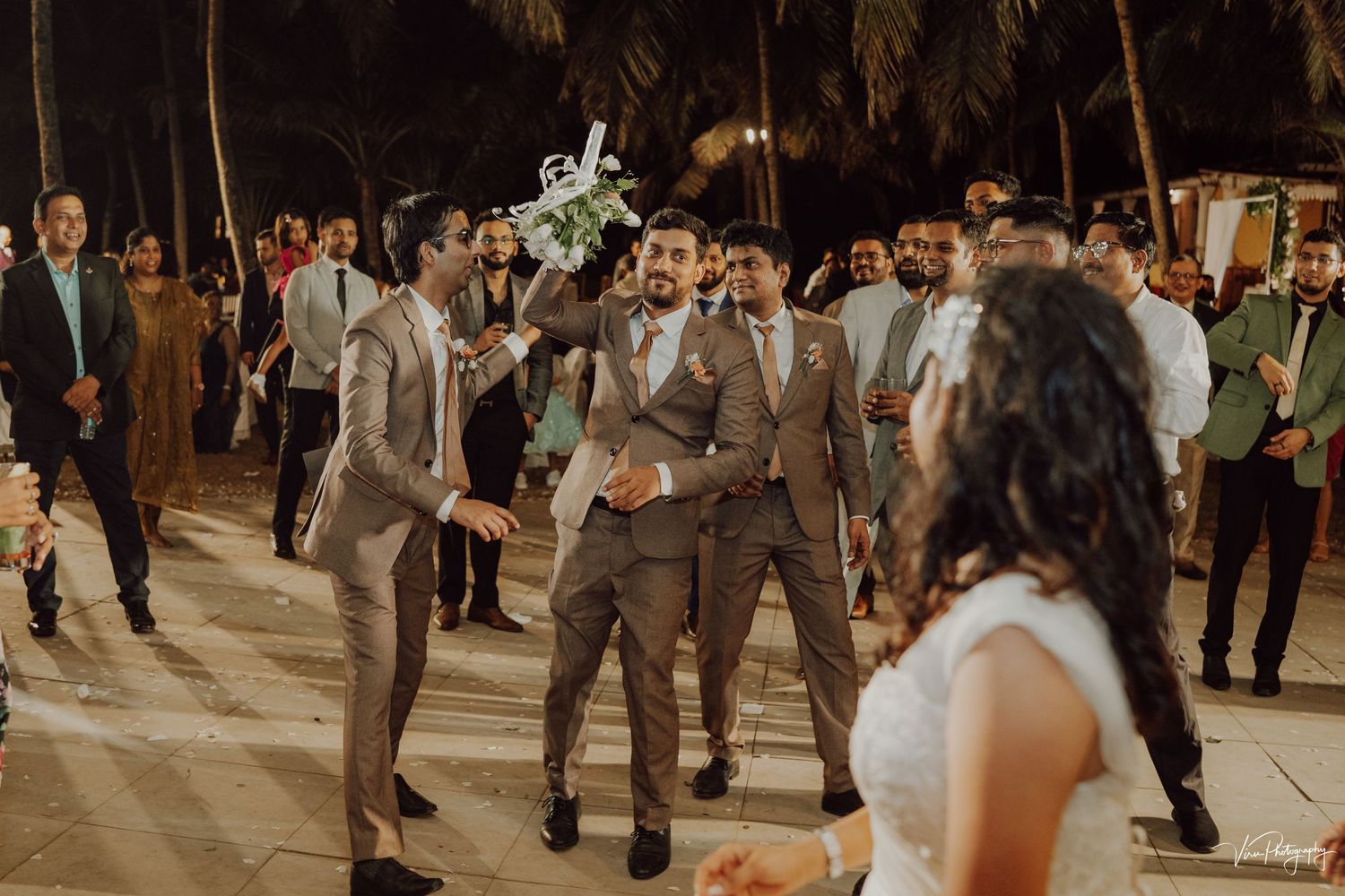 Wedding guests dance and celebrate on an outdoor palm-lined dance floor during an evening reception.