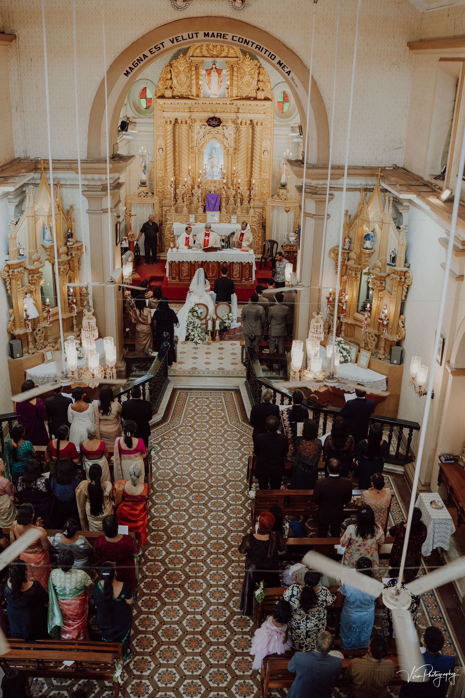 Aerial view of a traditional Catholic church wedding ceremony with ornate golden altar and guests seated in wooden pews.