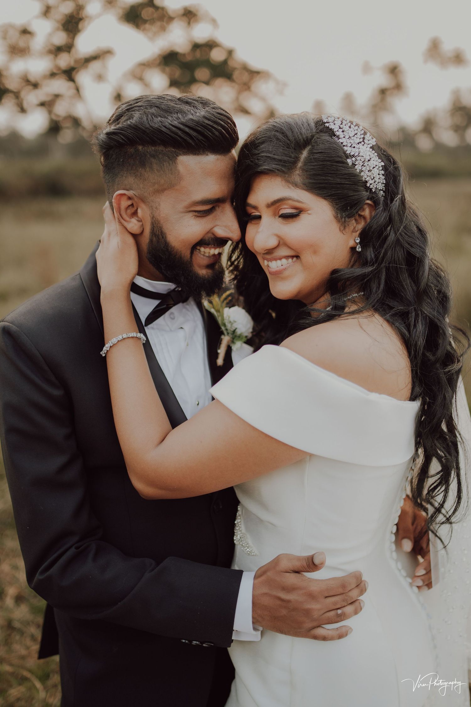 A romantic wedding portrait showing a couple embracing outdoors, with the bride wearing an off-shoulder gown and crystal tiara.