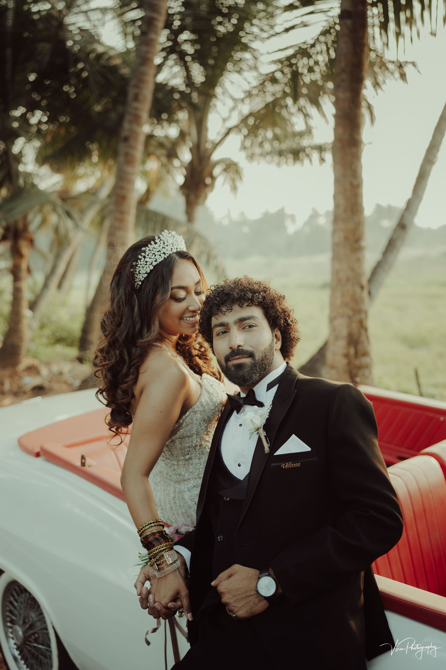 Stylish couple poses near vintage red car against tropical palm trees during golden hour wedding photoshoot.