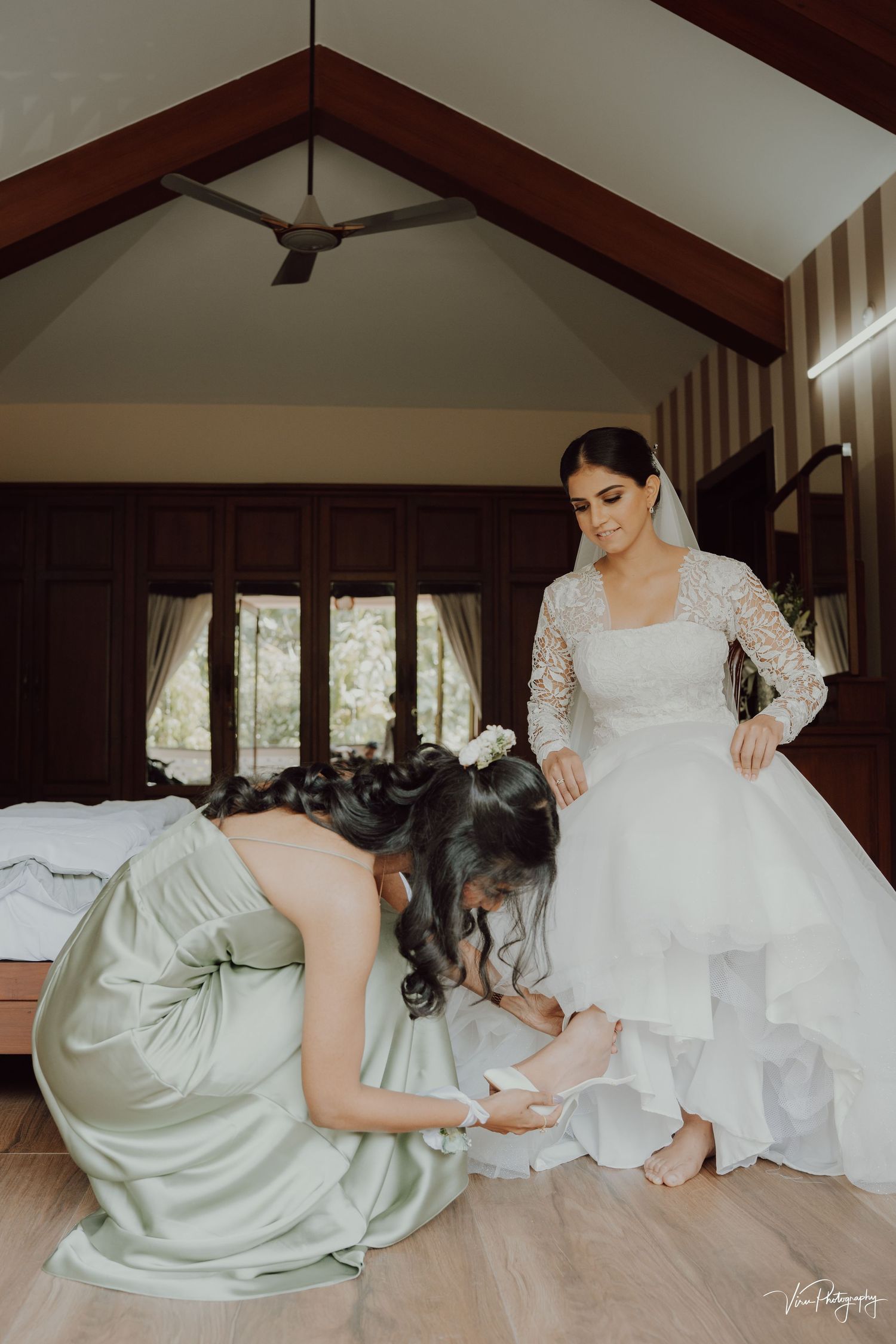 A bridesmaid in sage green dress helps adjust the hem of an elegant white wedding gown in a wooden interior room.
