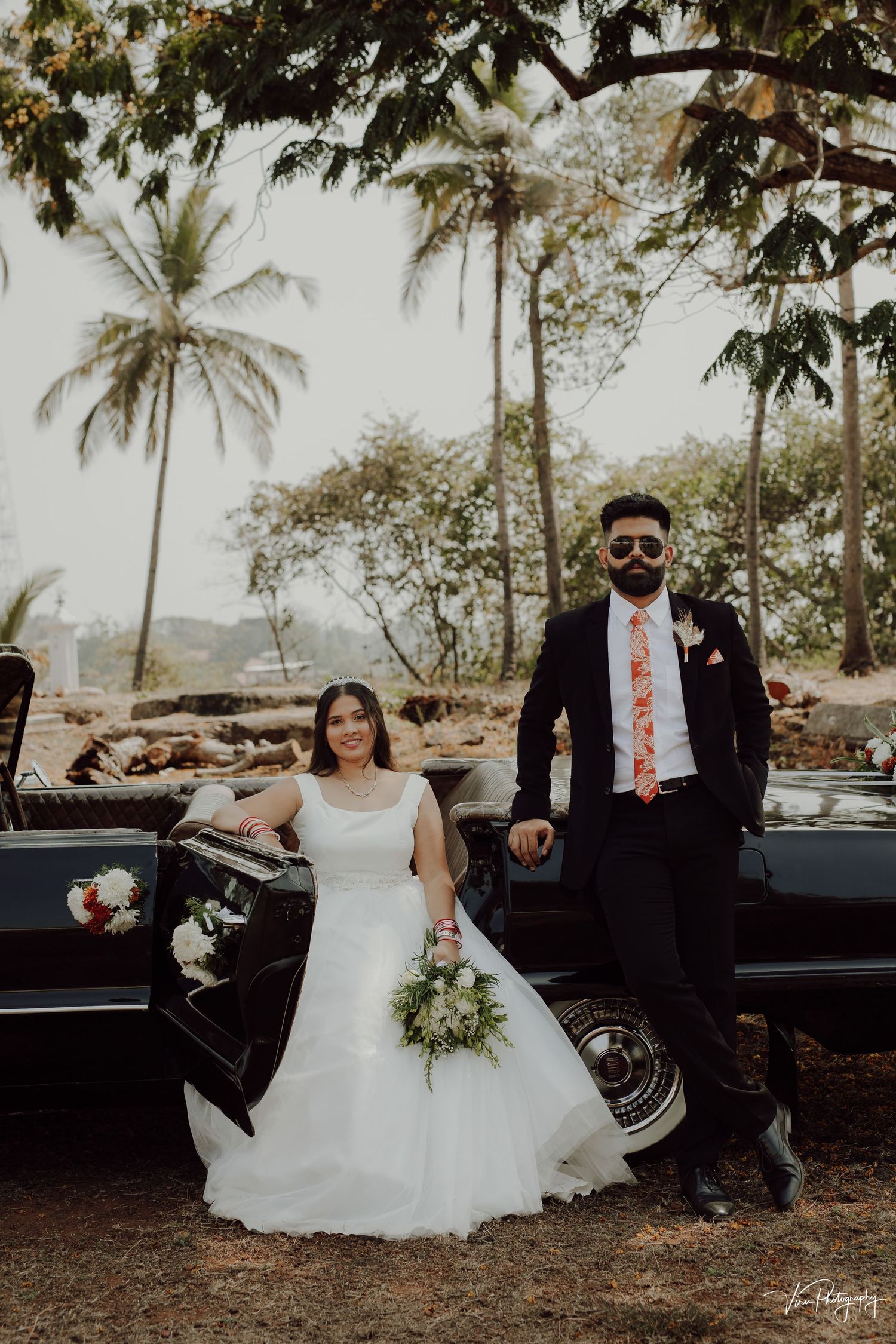 Newlywed couple poses next to vintage black car with palm trees and tropical scenery in background.