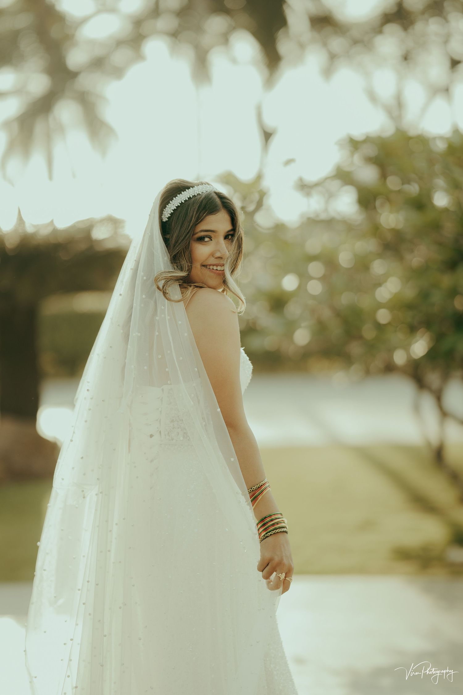 A bride in a flowing white wedding dress and veil poses outdoors with ethereal bokeh lighting in the background.