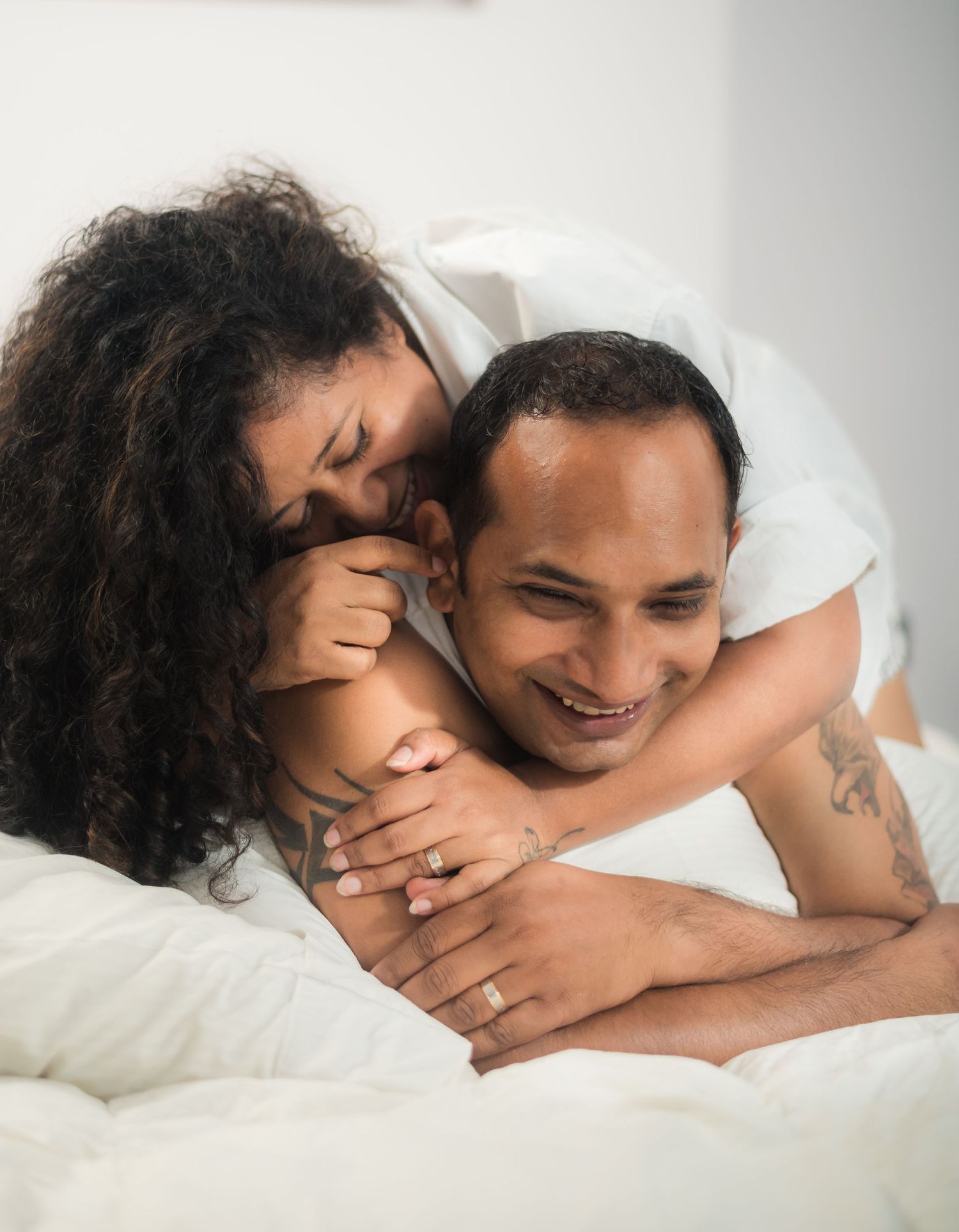A couple sharing an intimate moment while laying together in white bedding, showing warmth and affection.