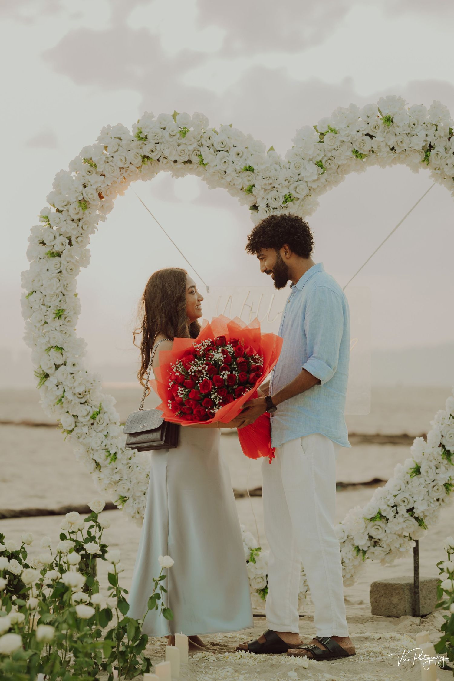 A couple stands beneath a white floral arch holding red roses during a romantic beach proposal.