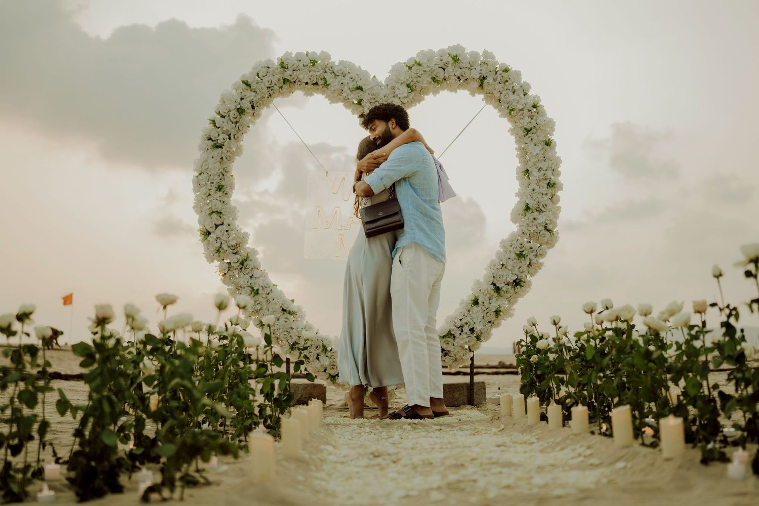 Romantic beach proposal setup with heart-shaped floral arch and embracing couple at sunset.