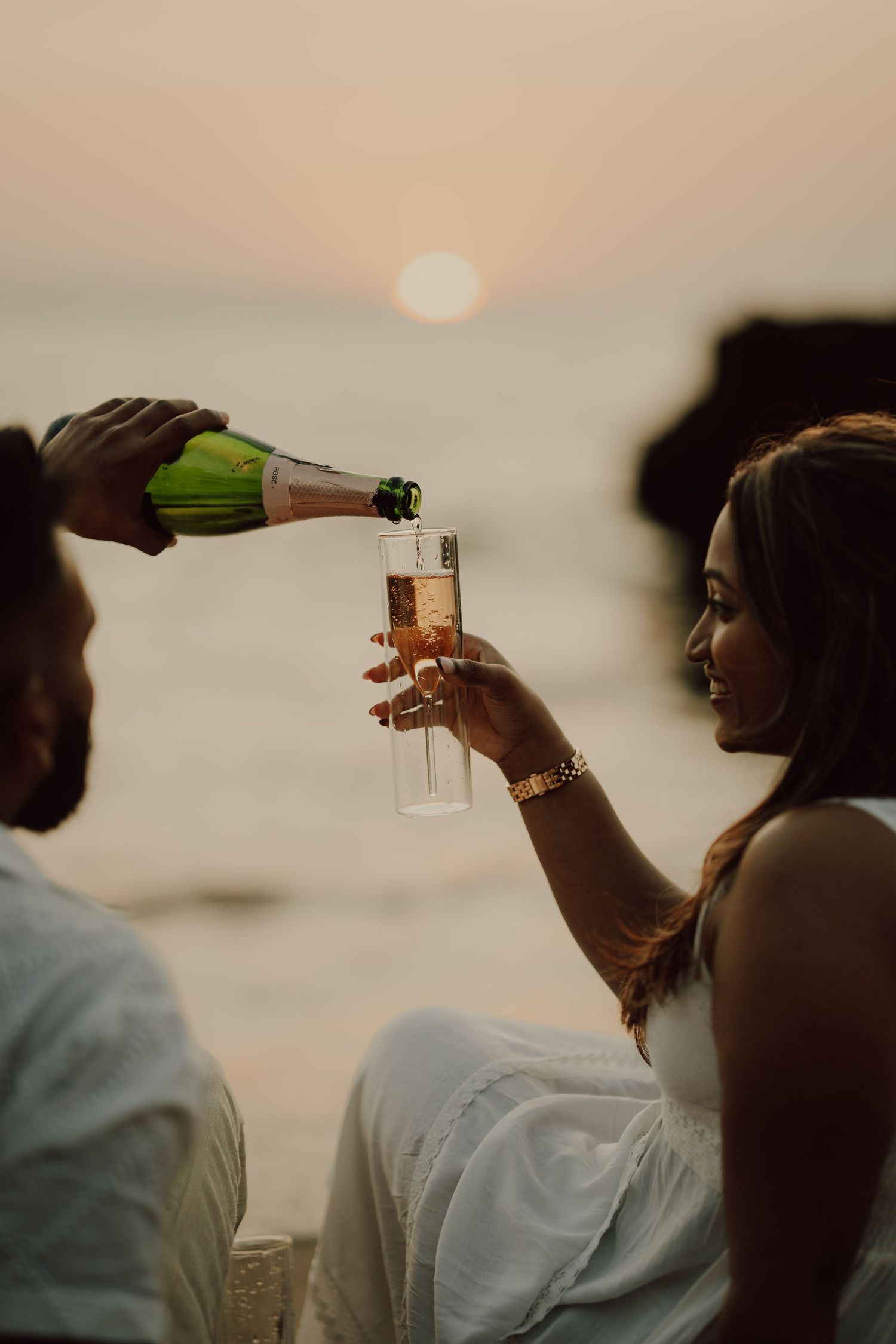 Two people in white shirts pour champagne during sunset at the beach.