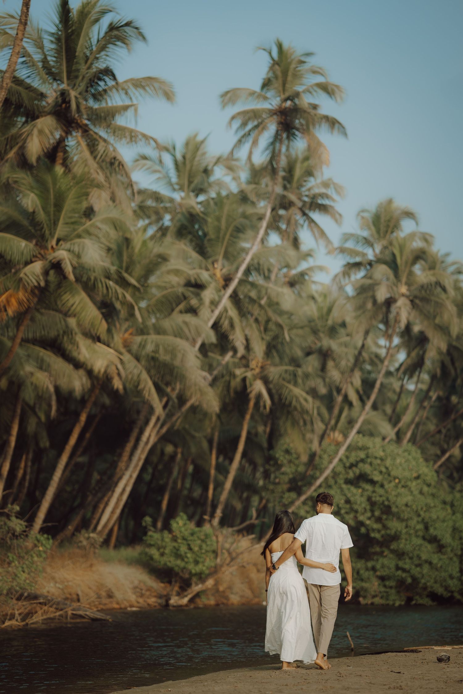 Couple walking along a tropical beach lined with tall palm trees and lush vegetation beside calm waters.