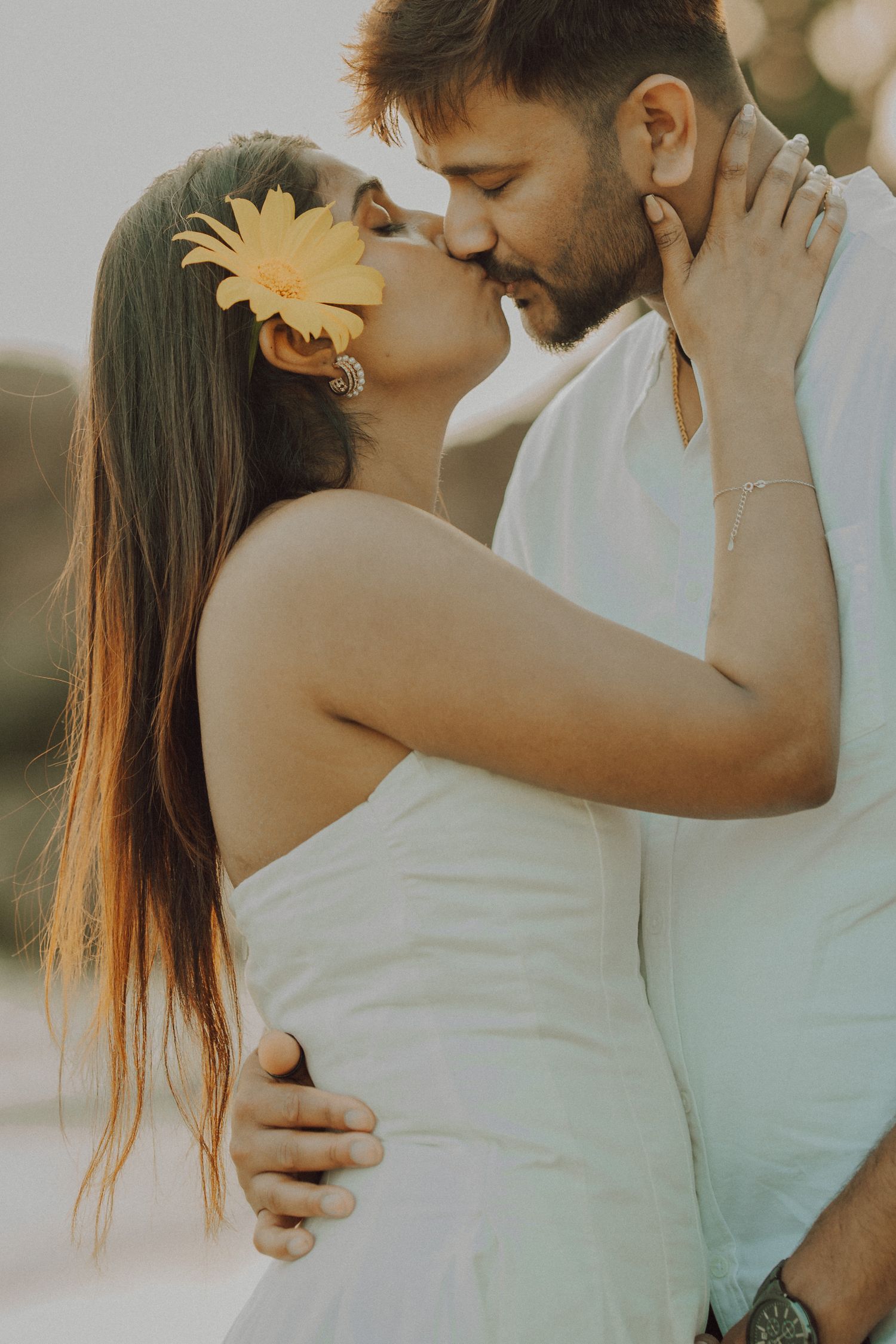 A romantic couple embraces in soft sunlight, with the woman wearing a white dress and yellow flower in her hair.