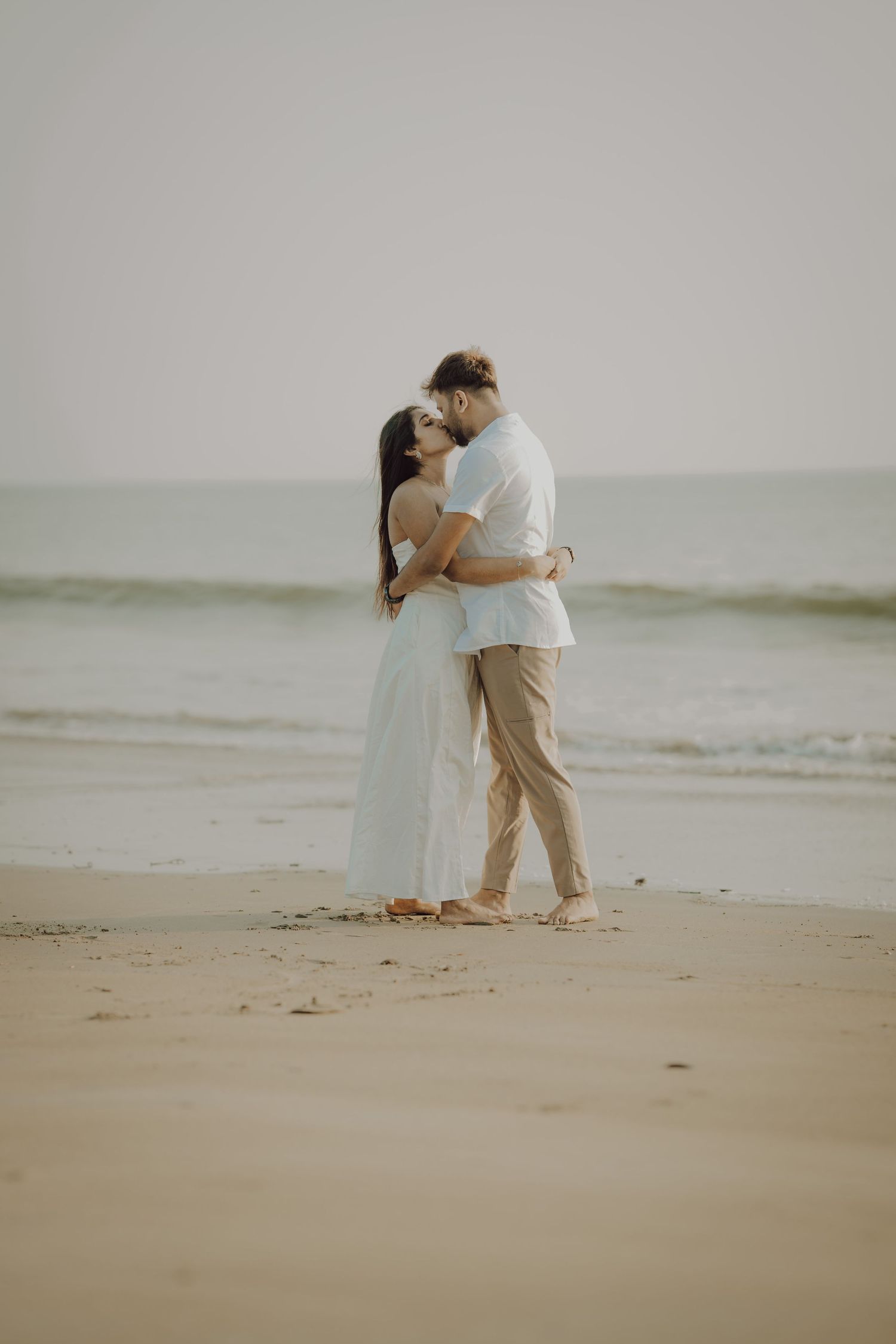 A romantic couple embraces on a misty beach shoreline wearing white and neutral colored outfits.