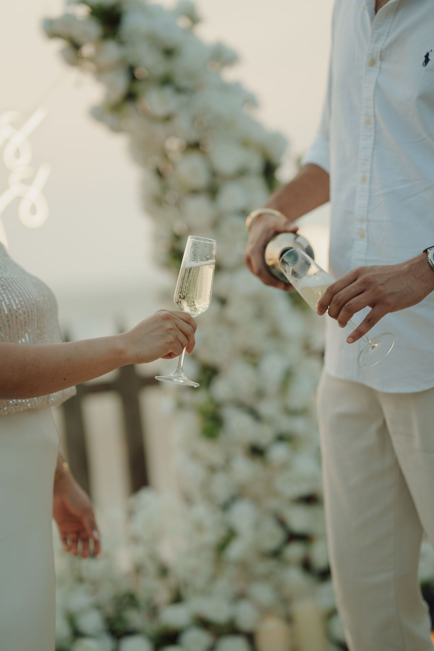 Couple celebrating with champagne at a white floral wedding arch, man pouring bubbly into elegant flute.