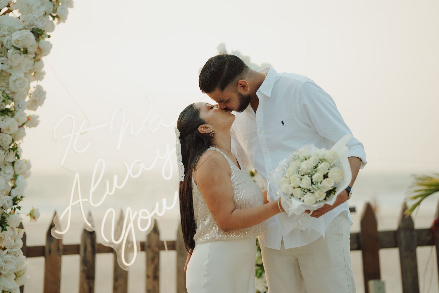 Couple sharing a romantic kiss at a beachside wedding ceremony with white floral arch and ocean backdrop.