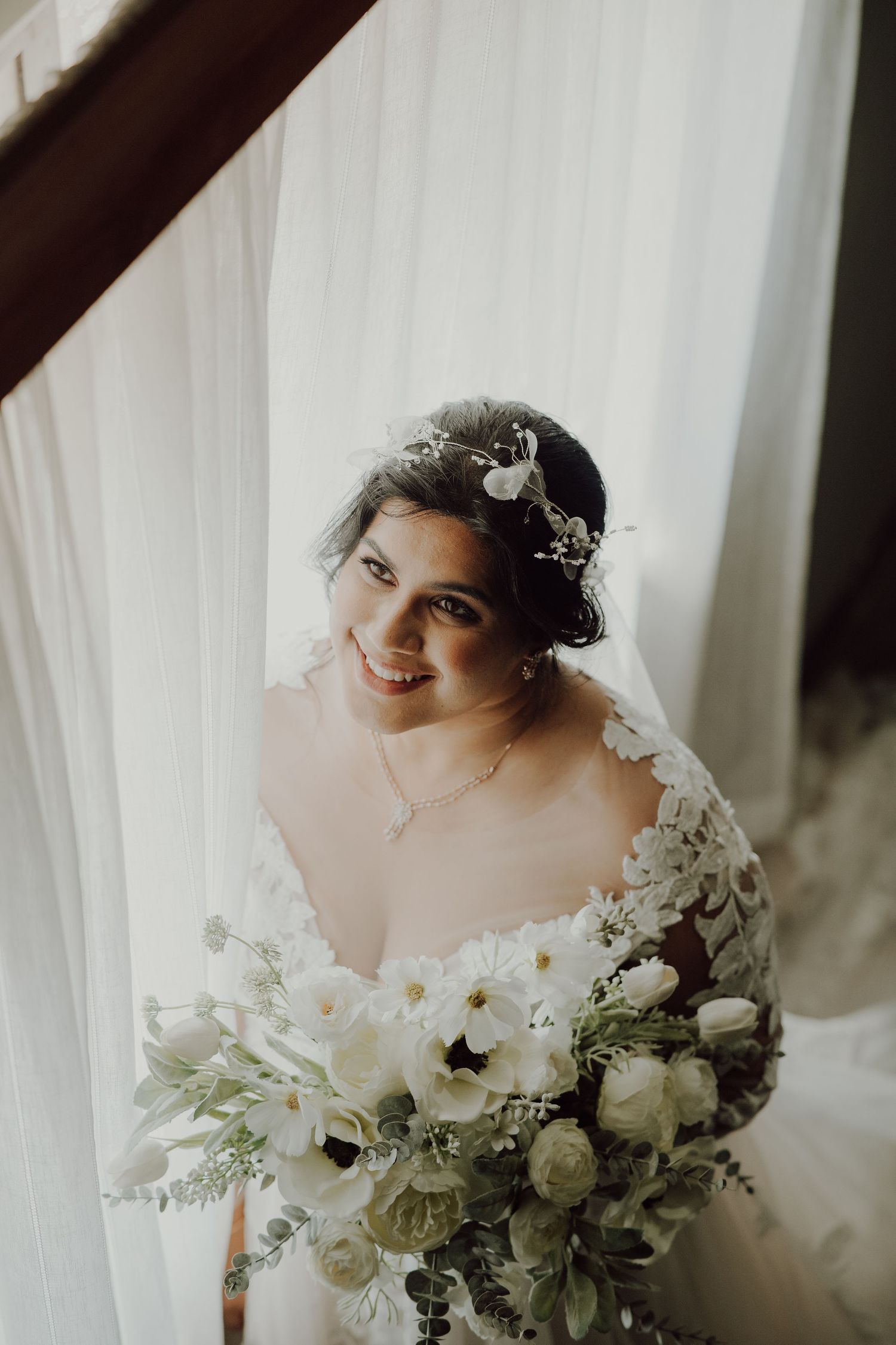 A bridal portrait with white floral bouquet and crystal crown against white curtains.