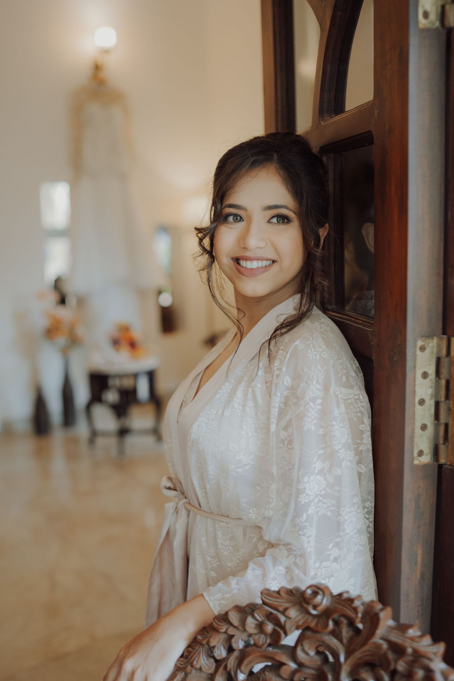 Smiling bride in a cream lace robe leans against a wooden door in a bright room with a hanging wedding gown behind her.