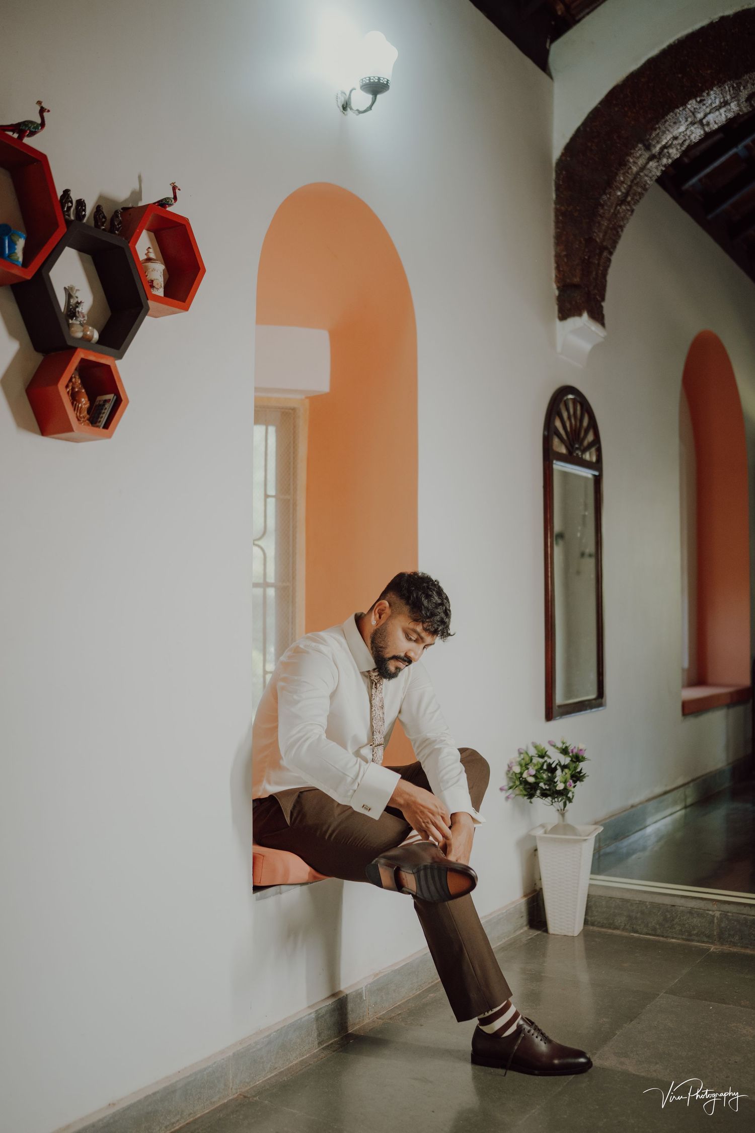 A person in formal attire sits in an elegant arched hallway with peach-colored walls and decorative hexagonal shelves.