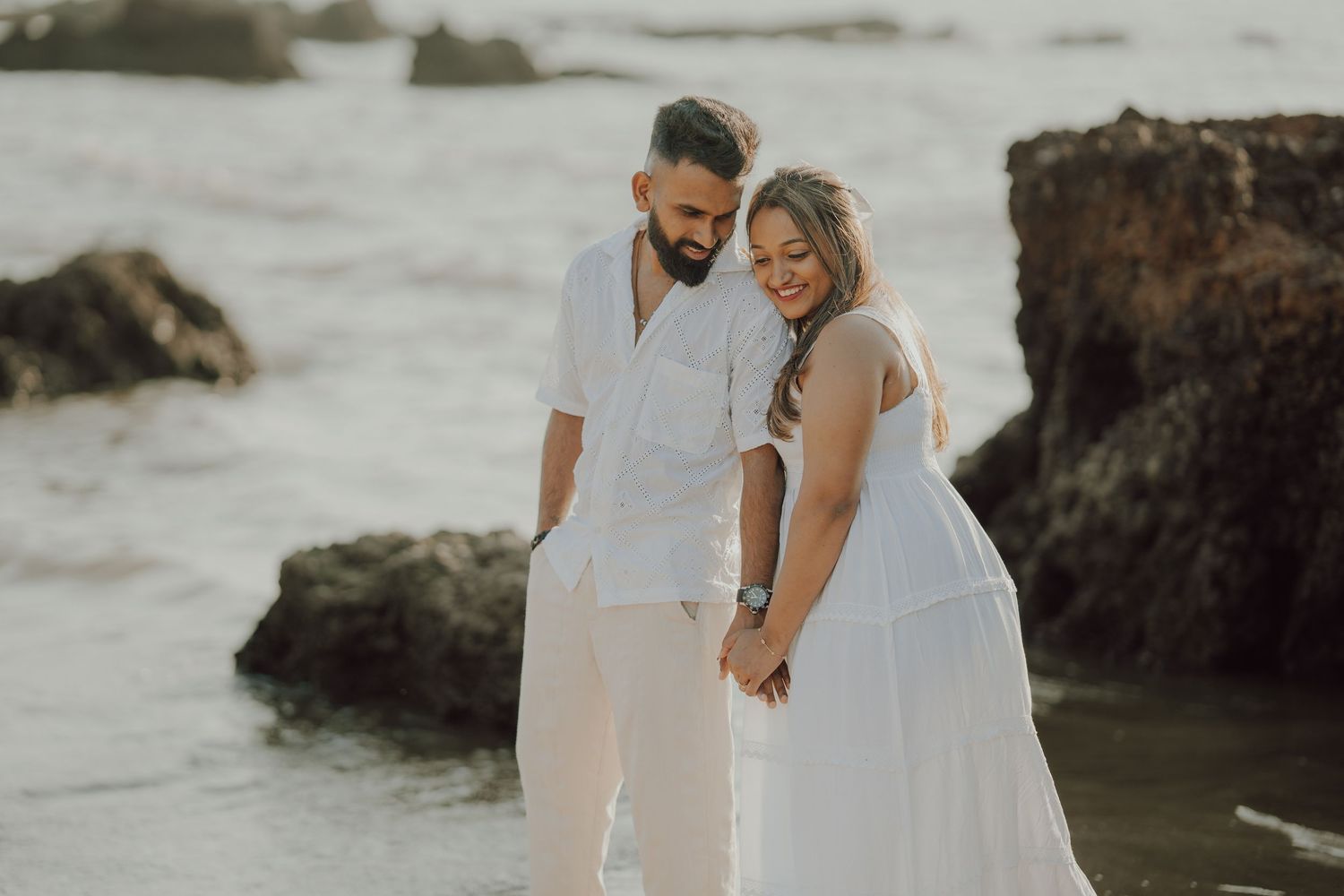 A couple in white wedding attire walks along a rocky beach during a romantic coastal photo session.