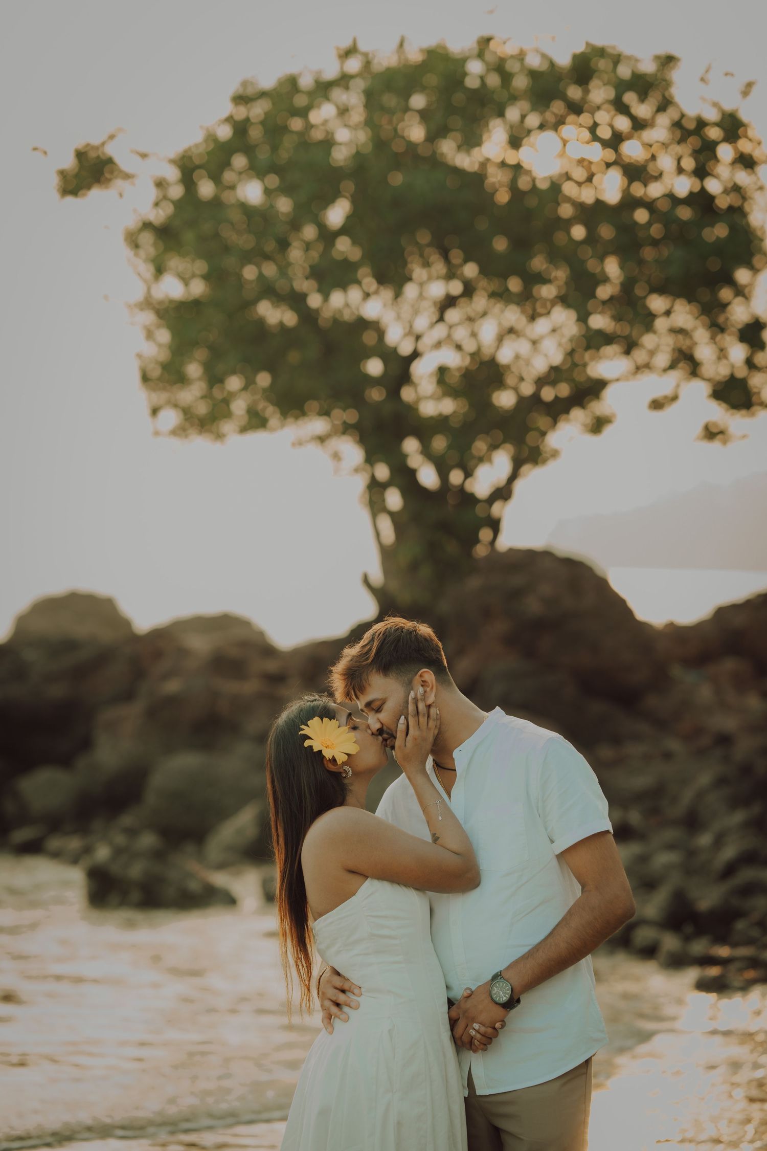 A couple shares an intimate moment while posing together against a dreamy sunset landscape with trees.