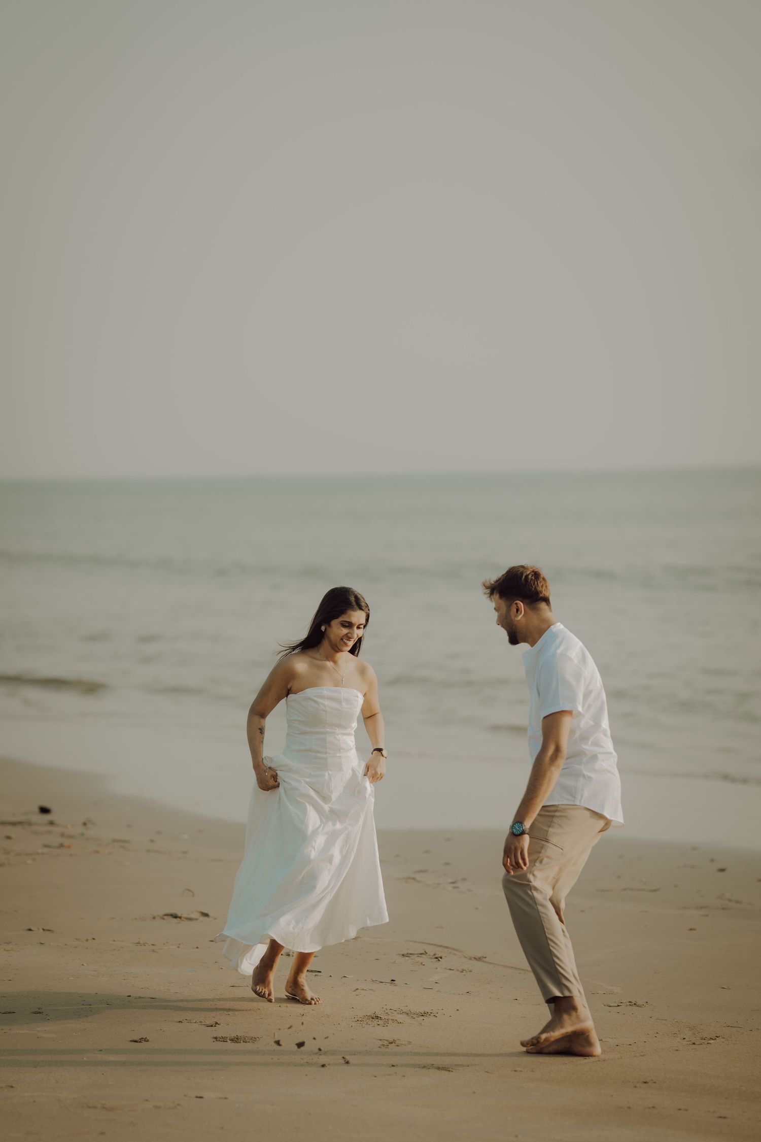 A couple in white and beige outfits playfully walks along a misty beach shoreline.