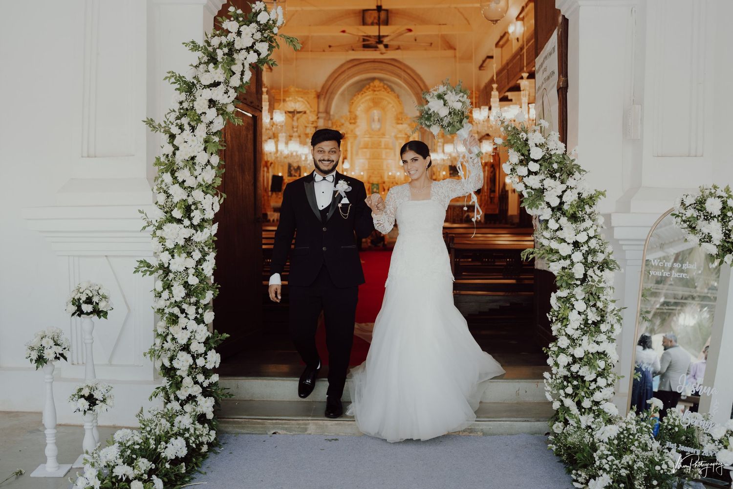 Two people exit a church through an elegant archway decorated with white floral arrangements and string lights.