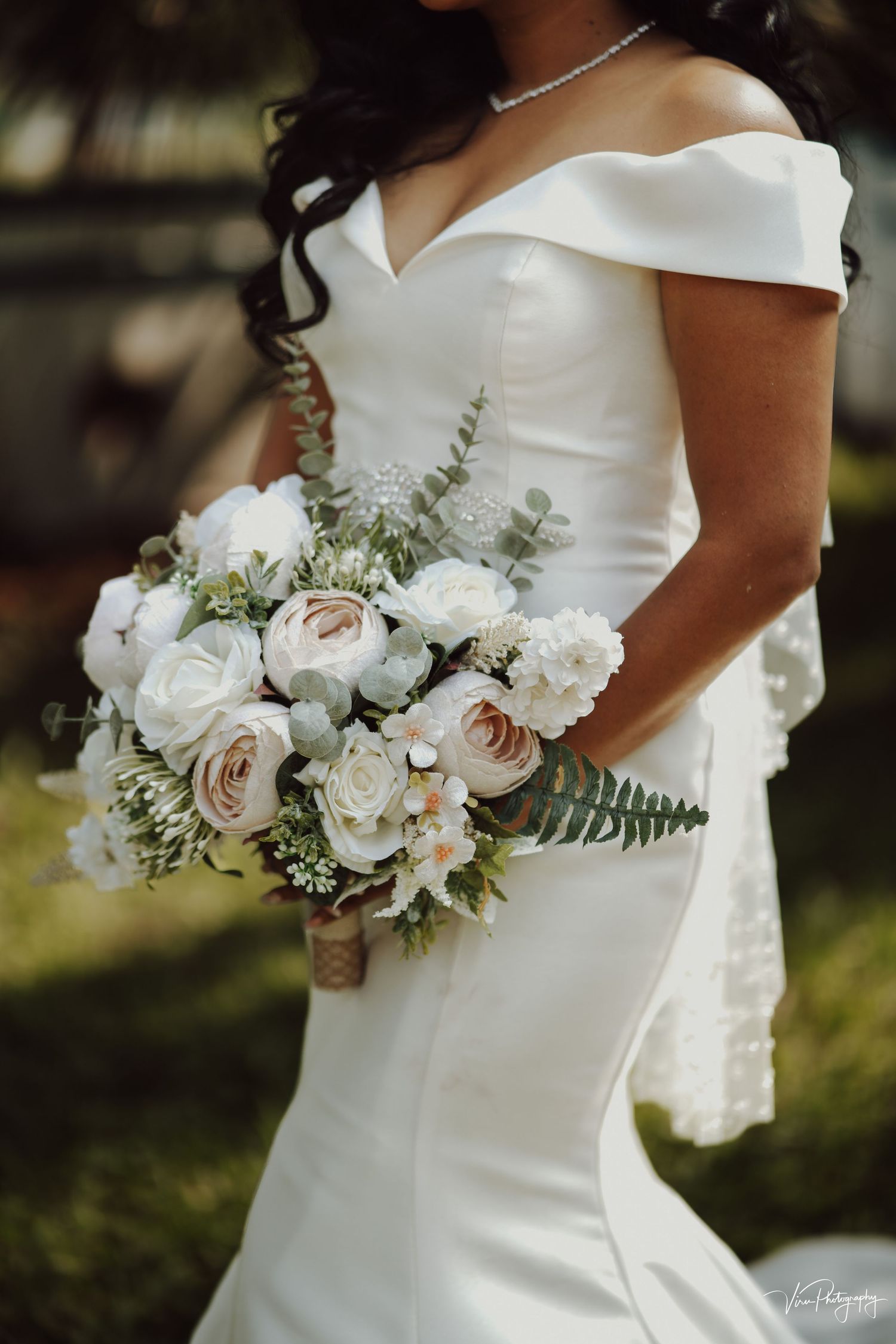 Close-up of elegant white mermaid wedding dress with off-shoulder neckline and blush rose bouquet with ferns.