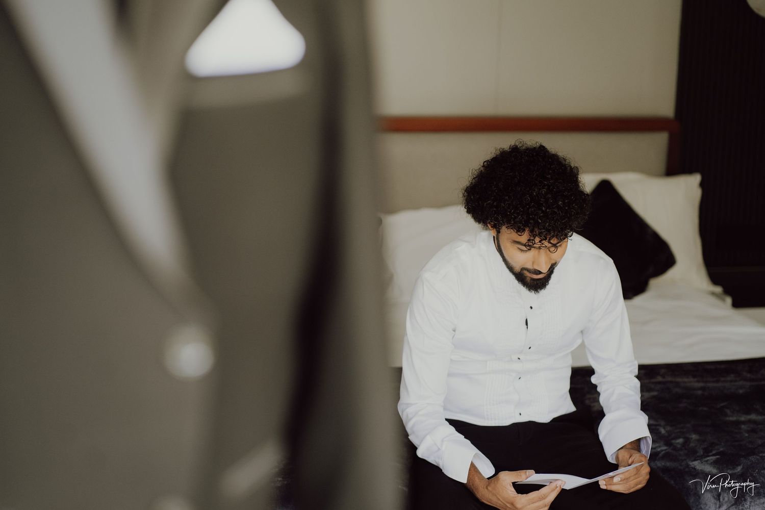 Person in a white shirt sits on edge of bed looking down at their phone in a dimly lit room.
