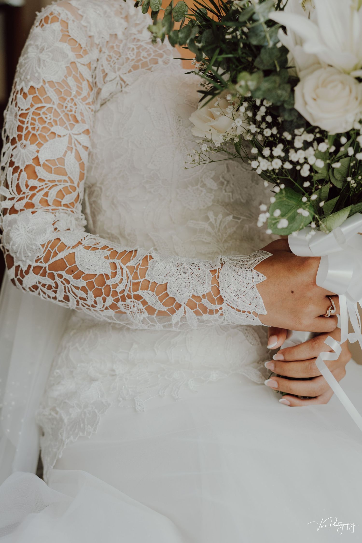 Close-up detail of white lace wedding dress sleeve and bouquet with roses and baby's breath flowers.